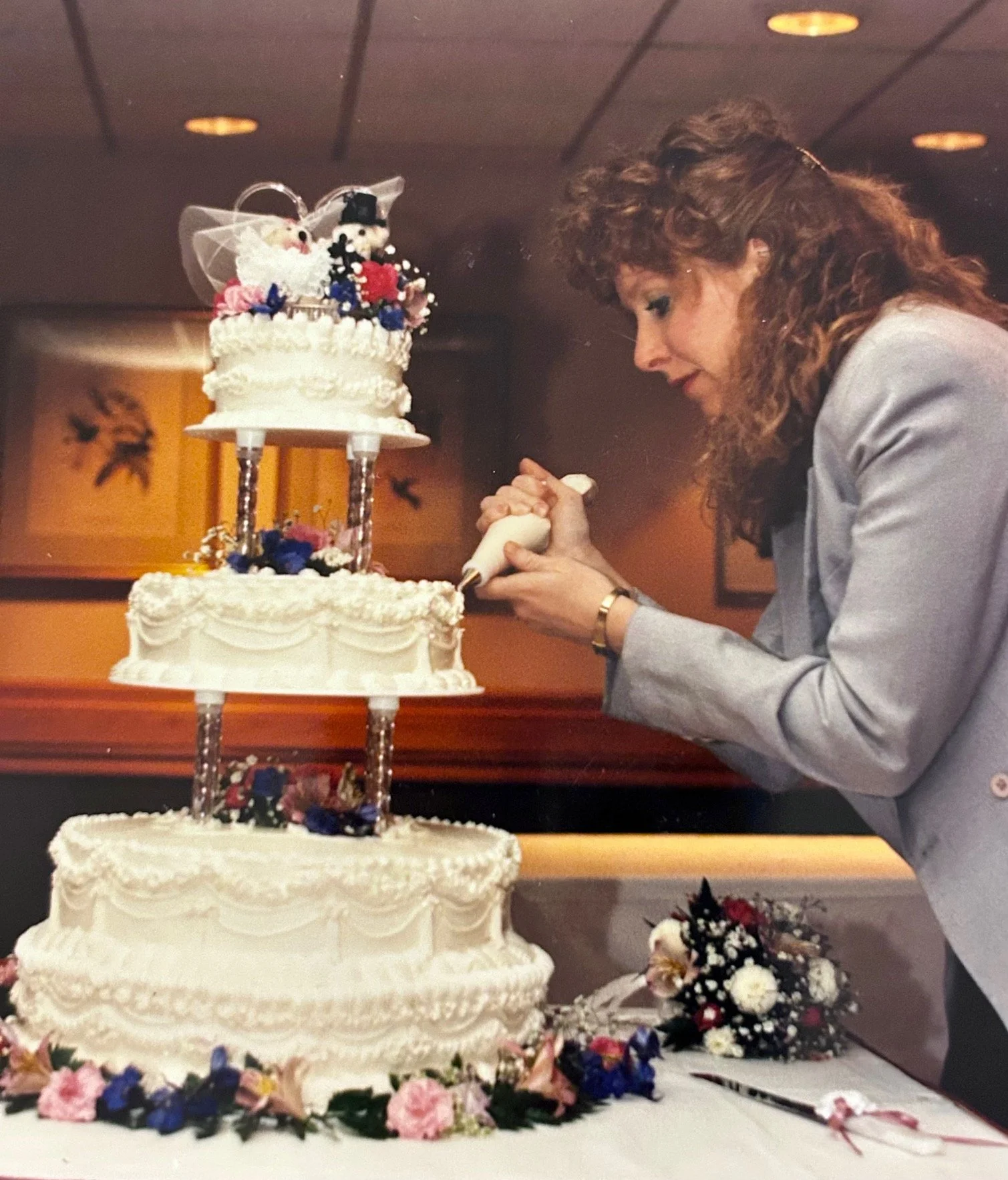 A woman with curly red hair, wearing a gray blazer, is holding a cake knife and preparing to cut a multi-tiered wedding cake decorated with white icing and flowers. The cake is on a table covered with flowers, and a small bouquet is on the table near the cake.