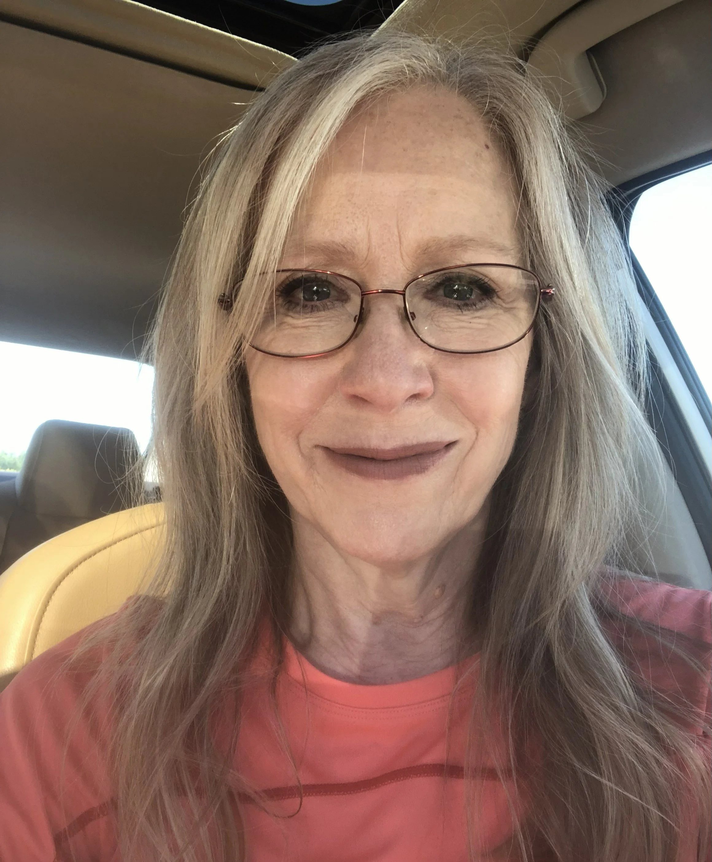 A woman with long blonde hair and glasses smiling inside a vehicle, wearing a pink shirt.