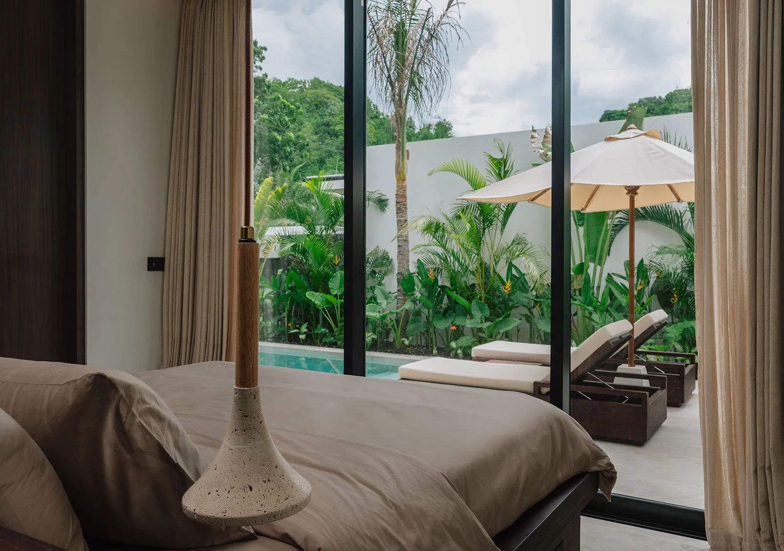 A bedroom with a large glass sliding door leading to a patio with lounge chairs, an umbrella, and lush greenery outside.