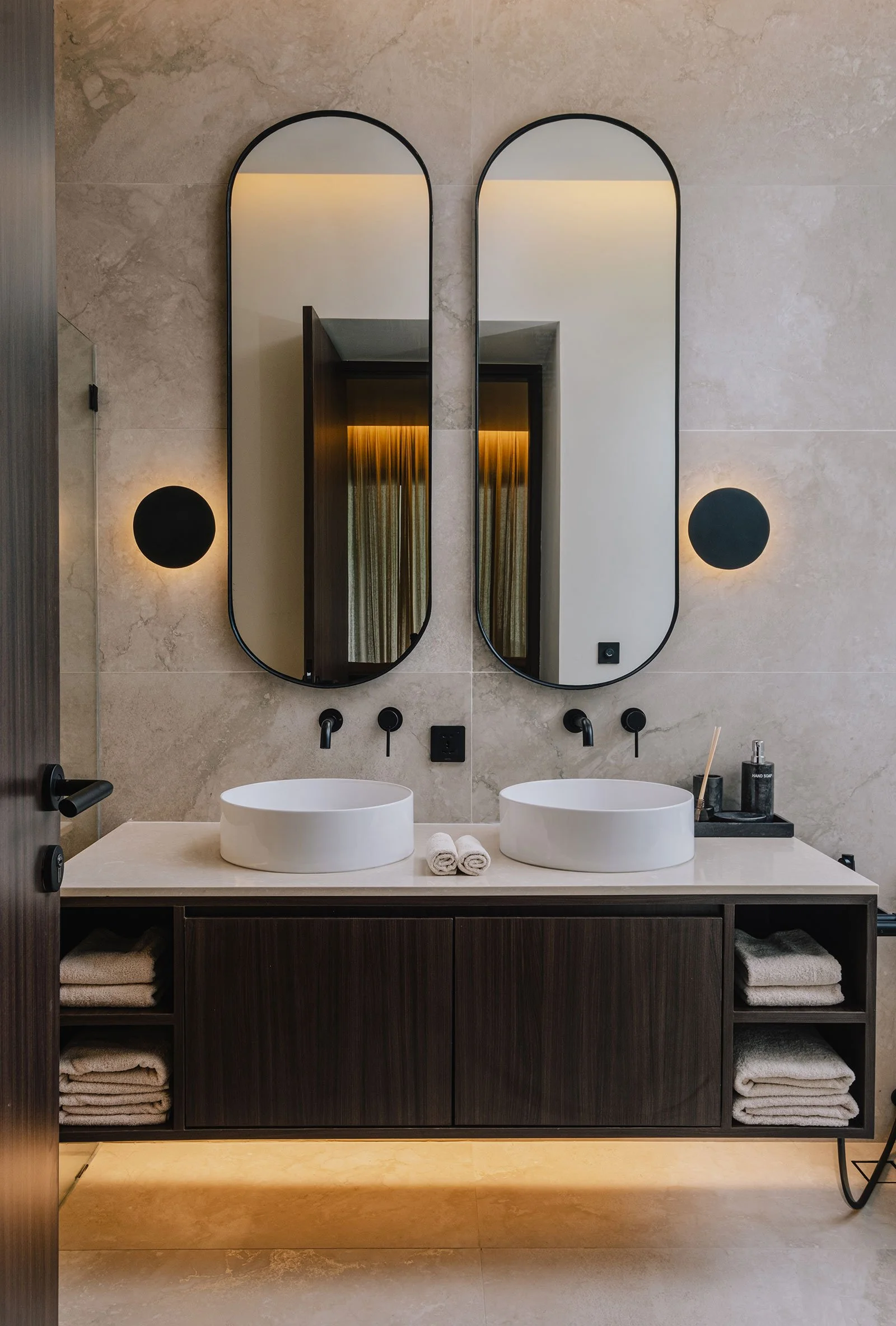Modern bathroom with two oval mirrors, black frames, and two white vessel sinks on a beige countertop with black faucets. Dark wooden cabinet below with open shelves containing folded towels. Wall-mounted black sconces and a soap dispenser on the rig