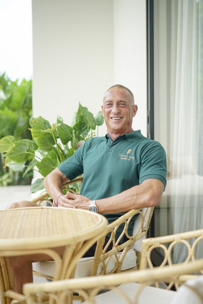 Smiling man sitting on a wicker patio chair on a balcony, with green plants and a window in the background.