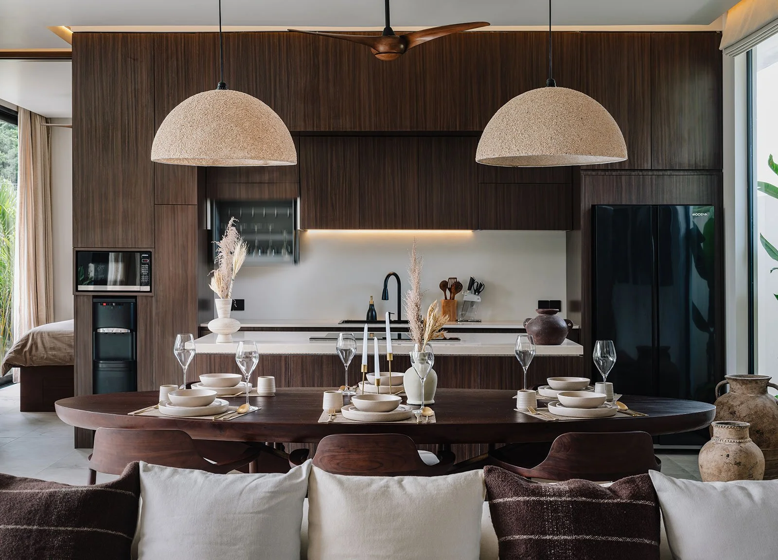Modern kitchen with wooden cabinetry and a dining table set with white plates, glasses, and gold utensils, decorated with beige vases and candles. Two large pendant lights hang above the table, and there are sliding glass doors to the outside.