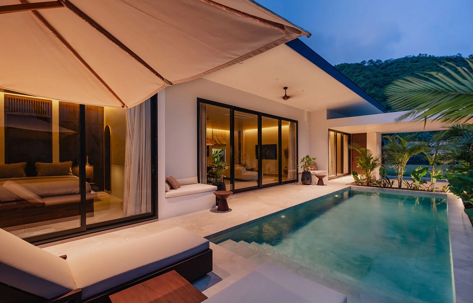 Modern outdoor patio with swimming pool, glass sliding doors to interior living space, lounge chairs, potted plants, and surrounding greenery at dusk