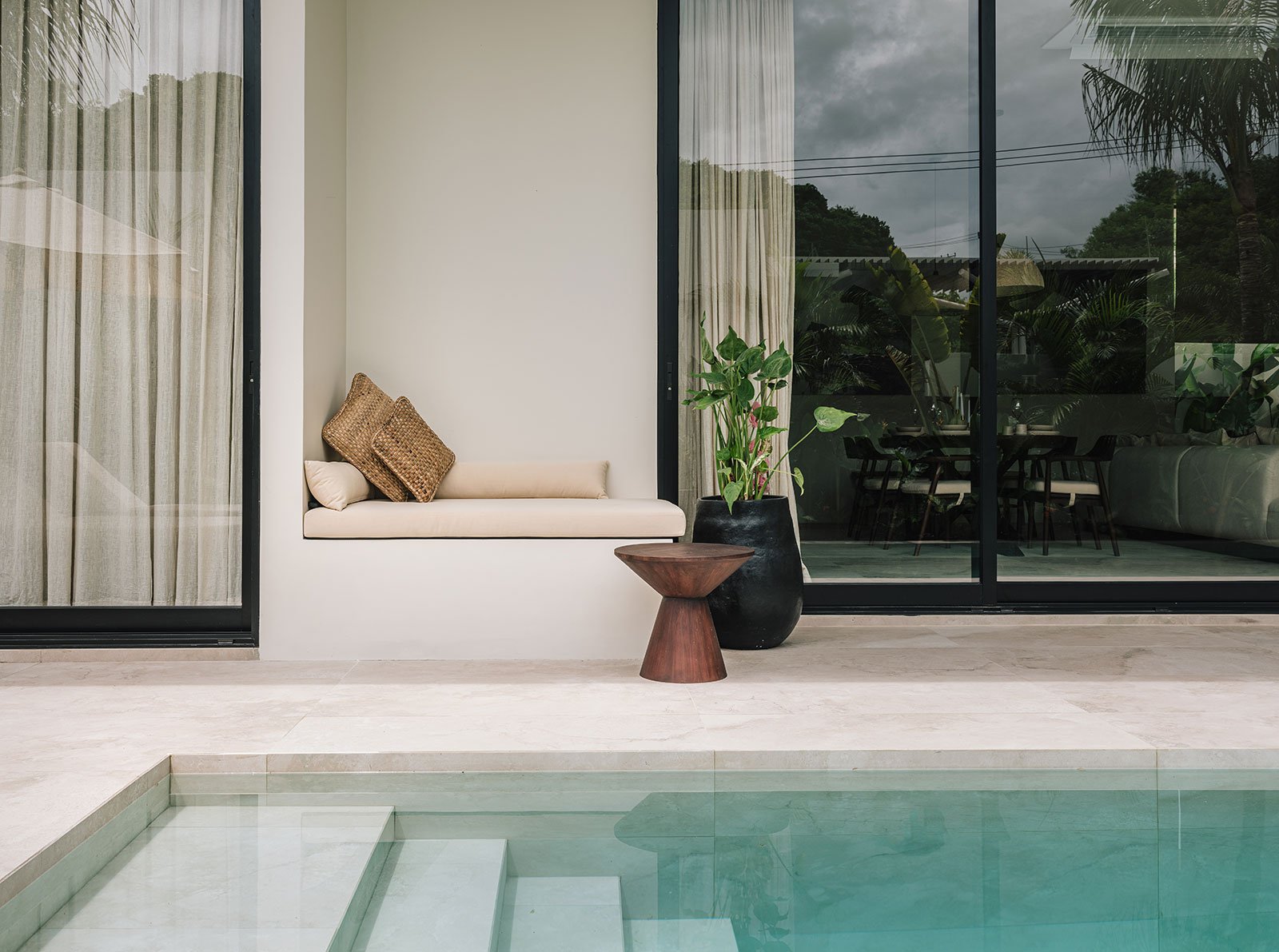 Modern indoor pool area with a built-in lounge seat, brown cushions, and a large potted plant. Glass sliding doors leading to an outdoor patio with plants and trees.