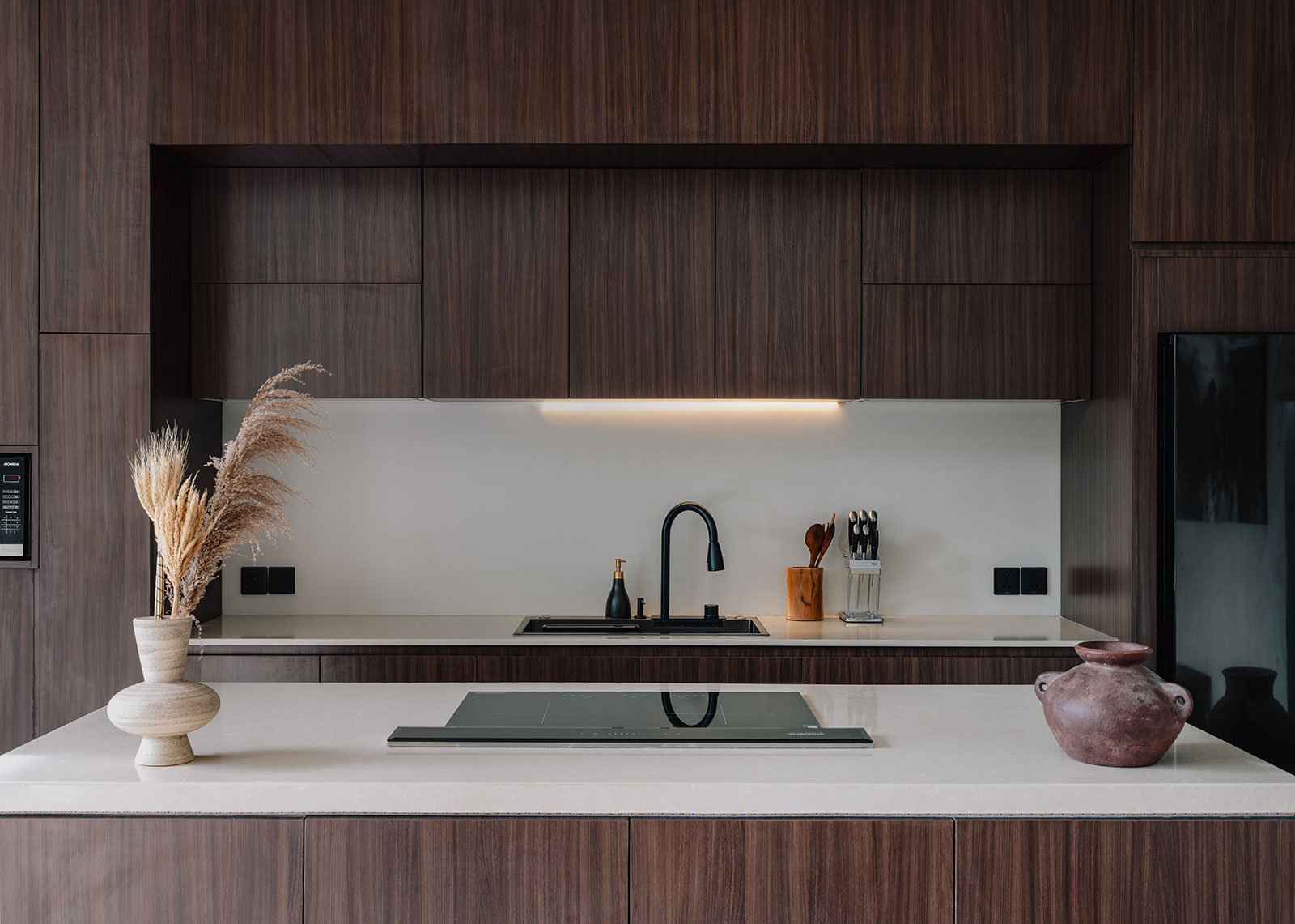 Modern kitchen with dark wood cabinets, white countertop, black sink faucet, decorative vase with dried plants, wooden utensils, and a pink pottery vase.