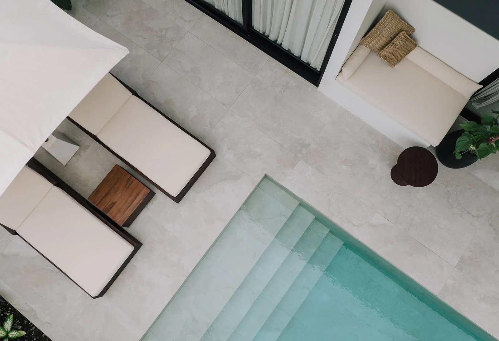 A modern interior space with tiled flooring, a glass staircase, a corner with a beige bench, a small wooden side table, a large potted plant, a window with curtains, and a beige slide with woven pillows.