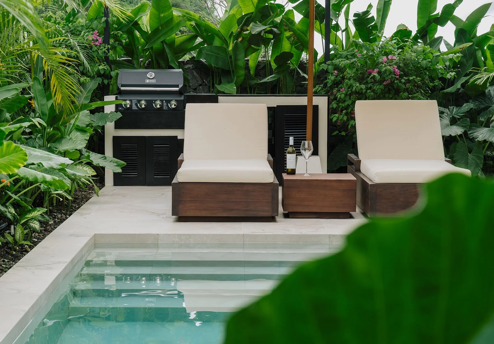 A backyard patio with two cream cushioned chairs, a small wooden side table holding a bottle of white wine and a glass, a barbecue grill, surrounded by lush green plants and foliage.