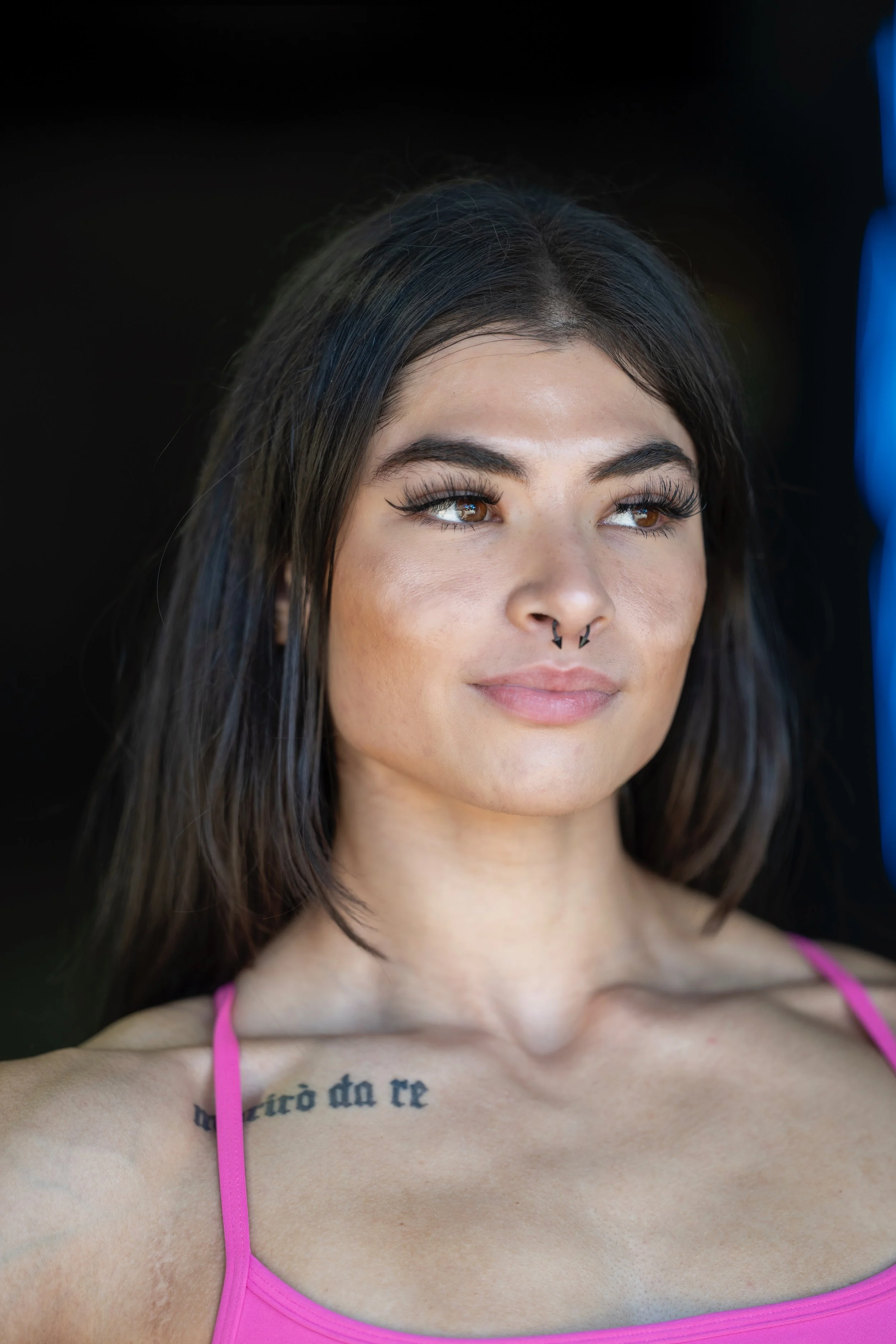 Close-up of a young woman with dark hair and brown eyes wearing a pink tank top, showing a tattoo on her chest.