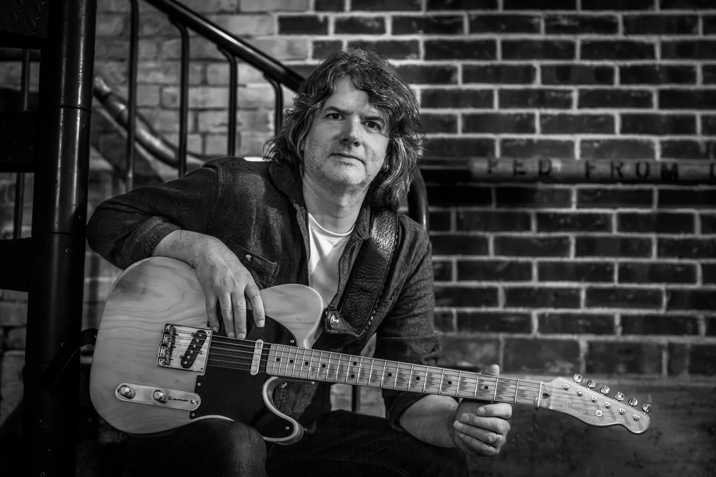 A man with medium-length wavy hair and a serious expression is sitting with a guitar against a brick wall background.