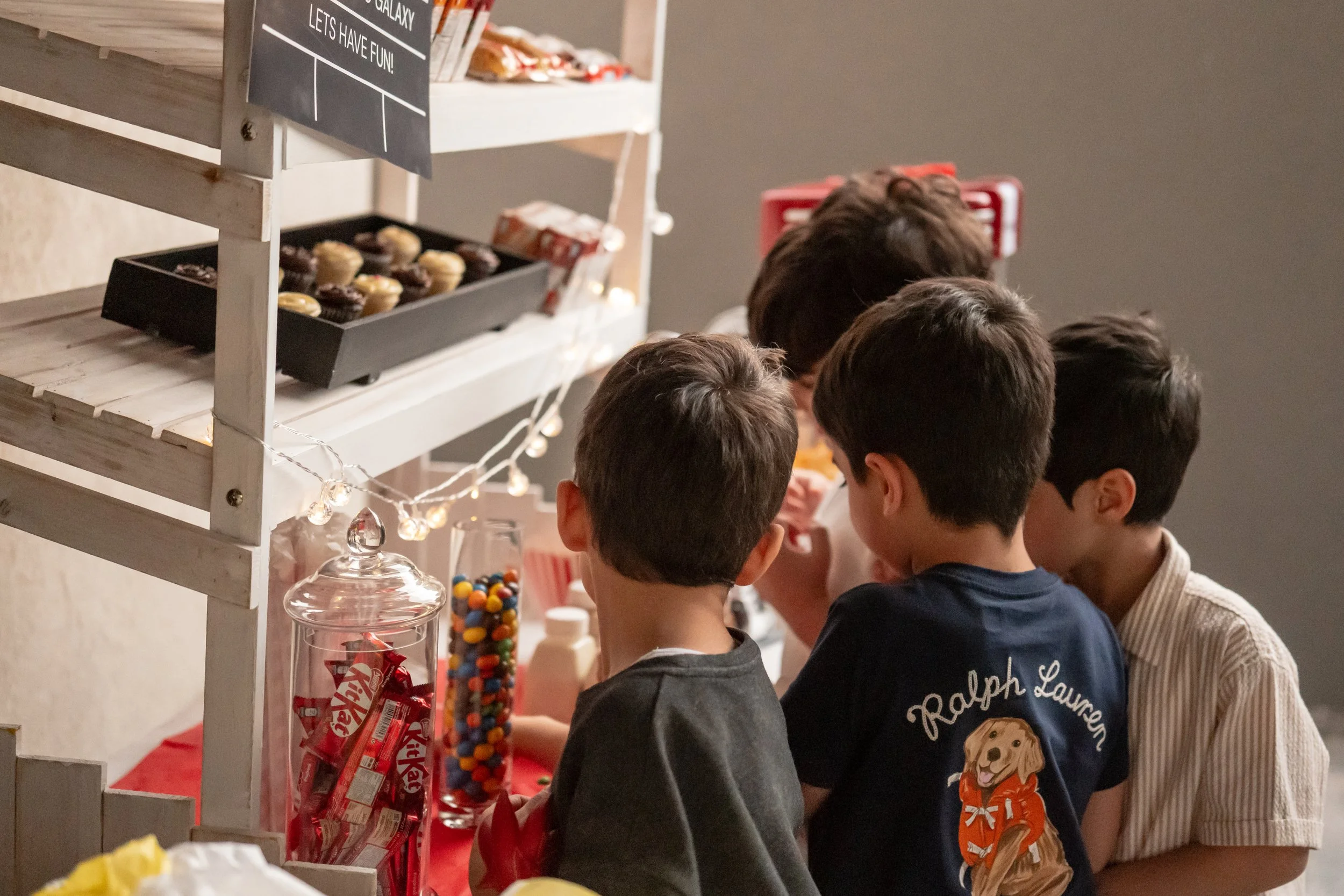 A group of children gathered around a candy table with various treats, including Kit Kat bars and colorful gumballs, at a party.