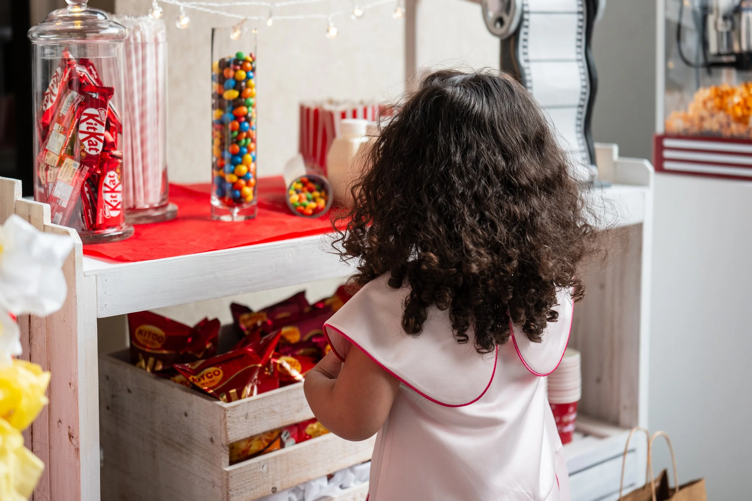 A young girl with curly hair wearing a white dress with pink trim stands in front of a snack stand, facing away. The stand has various candies including Kit Kat bars, Skittles, and other assorted sweets displayed in jars and baskets.