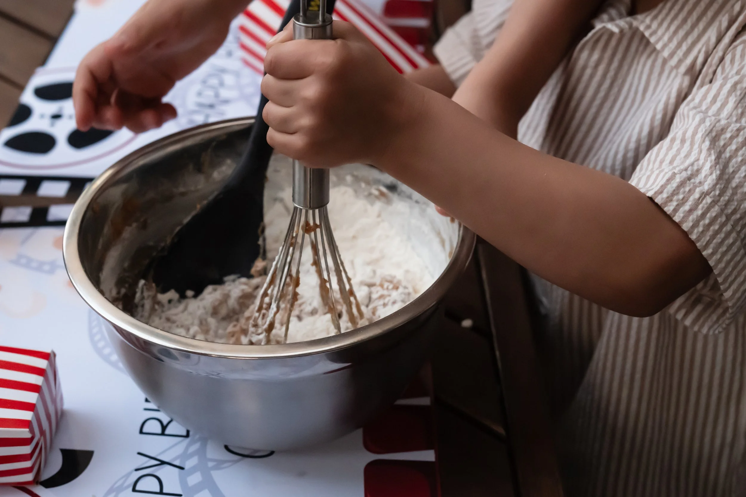 Child mixing cake batter in a metal bowl with a whisk, on a table decorated for a celebration.
