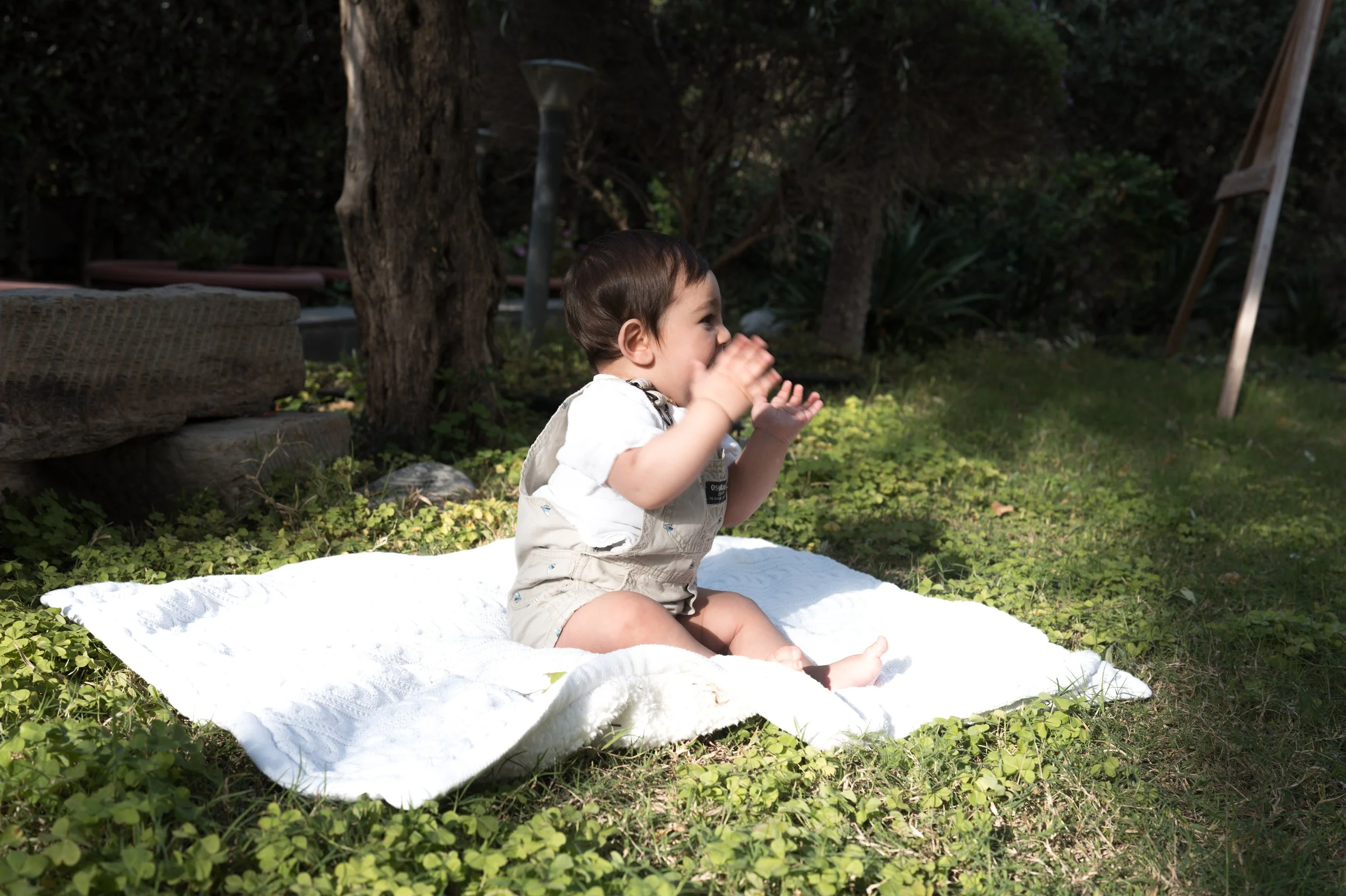 A young child sitting on a white blanket outdoors, clapping and smiling, surrounded by grass and trees.