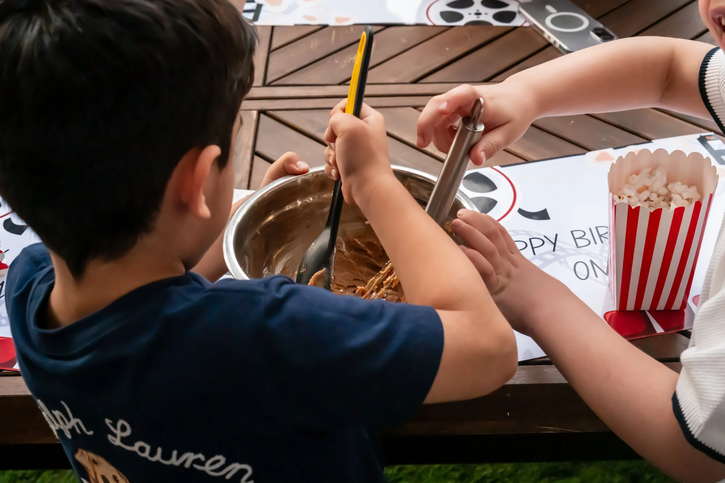 A boy and an adult stir ice cream in a metal bowl at a birthday party. There is a striped popcorn box and birthday decoration on the table.