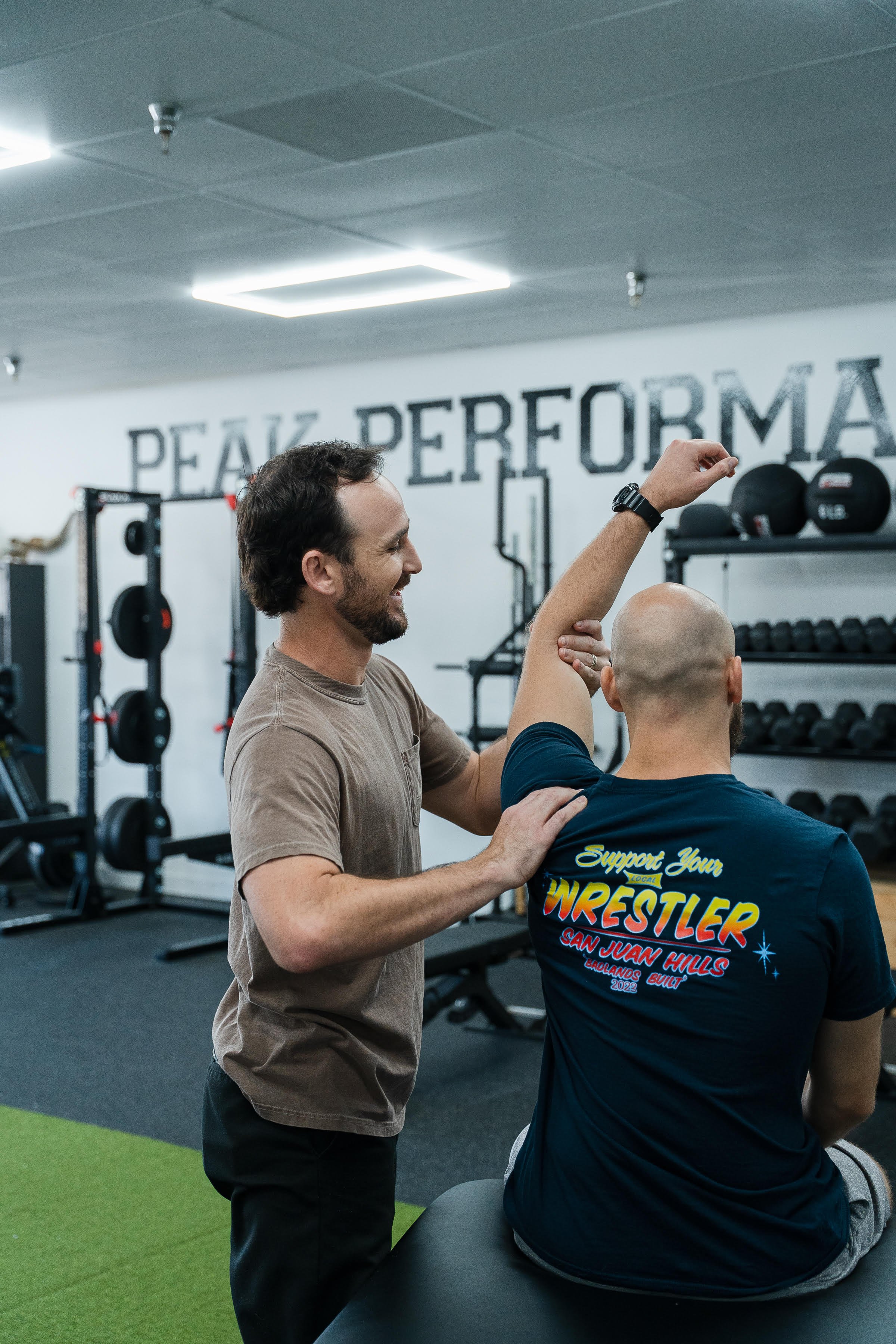 A man assisting a person with a shoulder stretch exercise in a gym. The background includes weight racks and the wall has the words 'PEAK PERFORMANCE' written on it.
