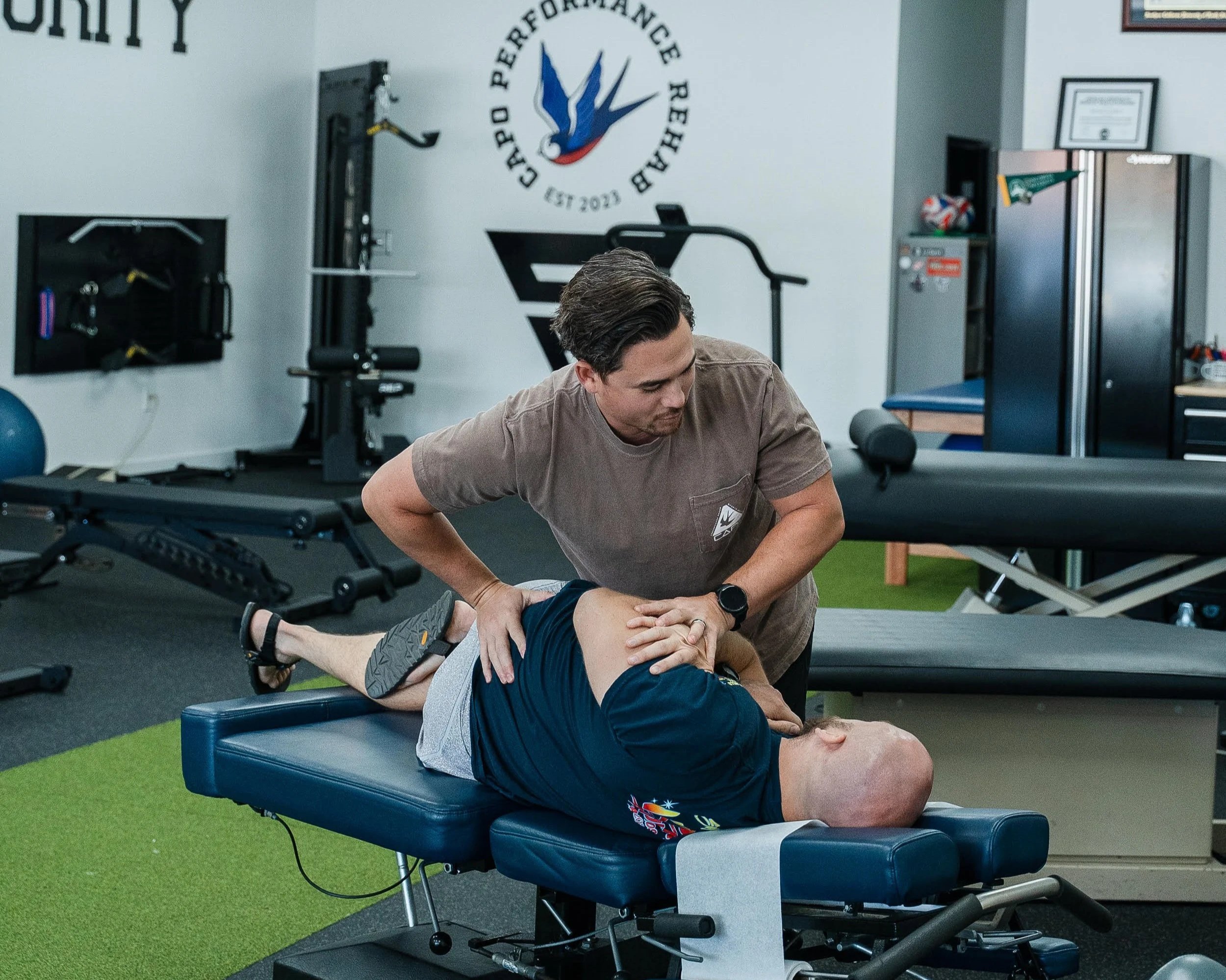 A physical therapist is performing a stretching exercise with a patient lying face up on a padded table in a fitness or rehabilitation center. The center has gym equipment and a logo on the wall that reads 'Society of Performance Rehabilitation,' established in 2023.