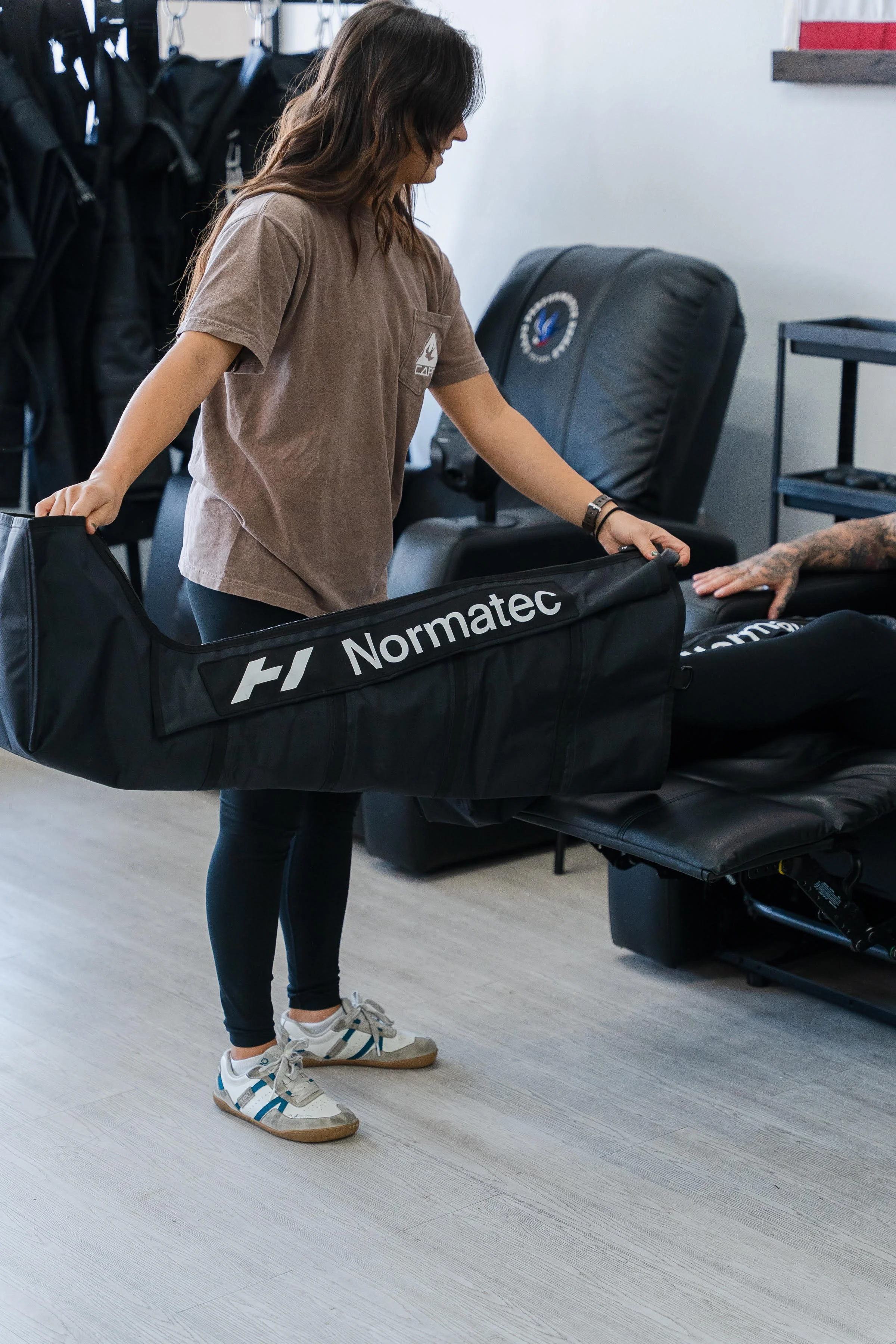 A woman with long brown hair wearing a brown t-shirt, black leggings, and white sneakers with blue accents, is lifting a leg compression boot labeled 'Normatec' in a room with a light wood floor, black chairs, and other compression hanging on hooks.