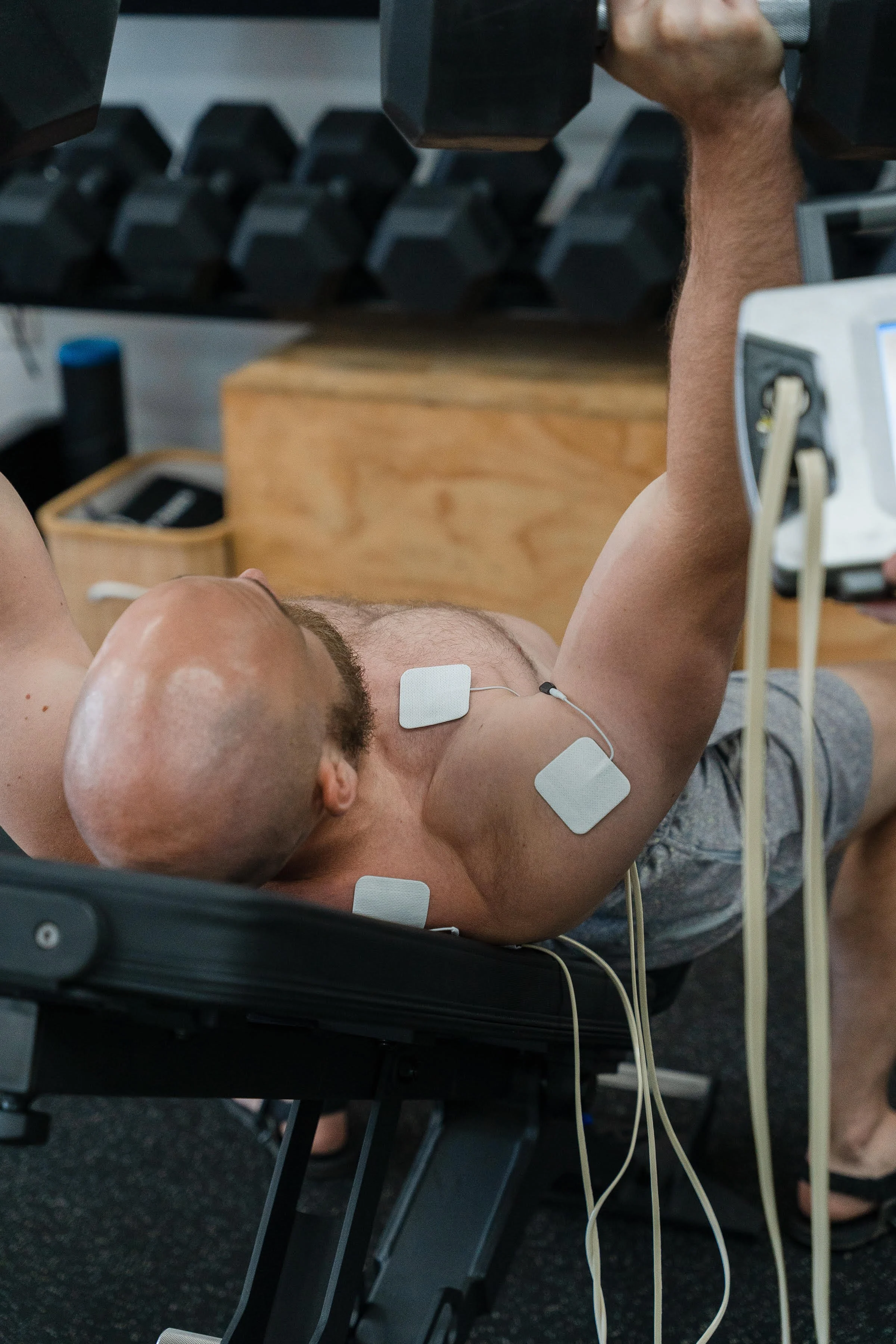 A man with a shaved head exercising at a gym, performing a dumbbell shoulder press while lying on a bench. He has a stim machine (ARP) attached to his chest and shoulder.
