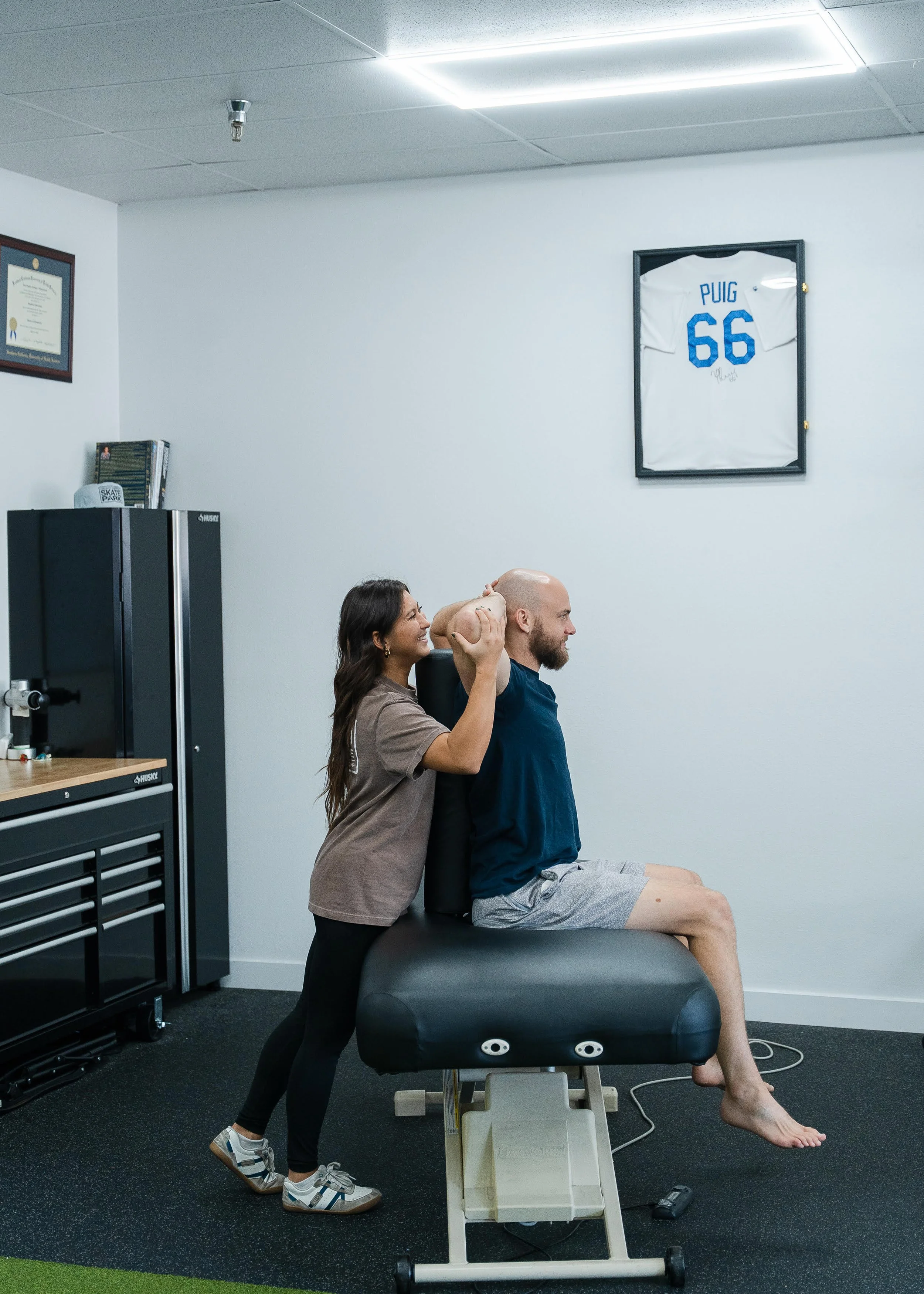 A physical therapist is performing a neck and shoulder stretching exercise with a male patient who is sitting on a treatment table. The therapist is standing behind him, gently supporting his head and neck. The patient is a young man with a bald head, wearing a navy blue shirt and gray shorts, seated with his eyes closed. The room has white walls, a framed sports jersey of Puig number 66 hanging on the wall, a black cabinet, and some exercise equipment.
