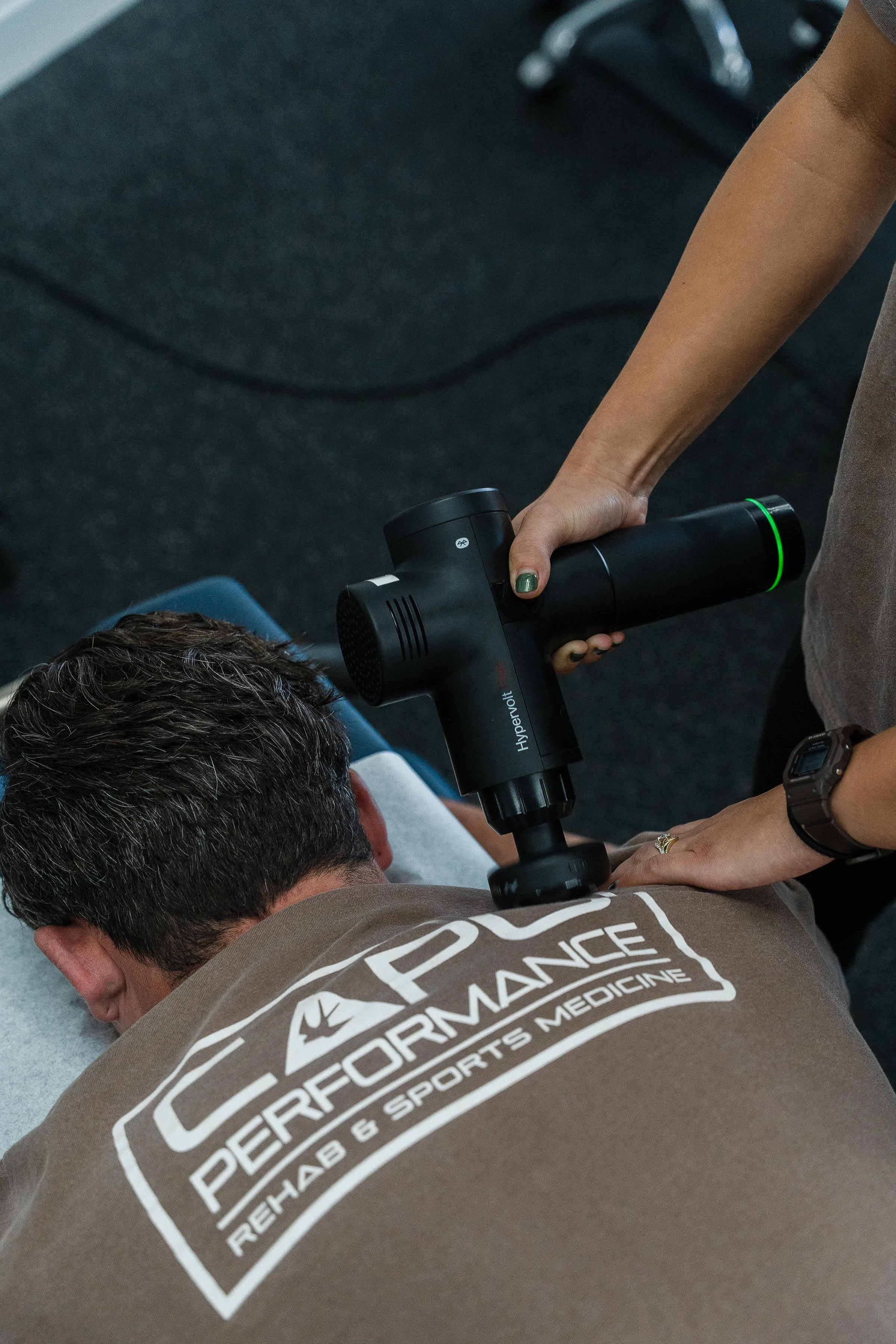 Person lying on the ground receiving a massage or therapy with a handheld device, wearing a shirt that reads 'Performance Healthcare & Sports Medicine.'