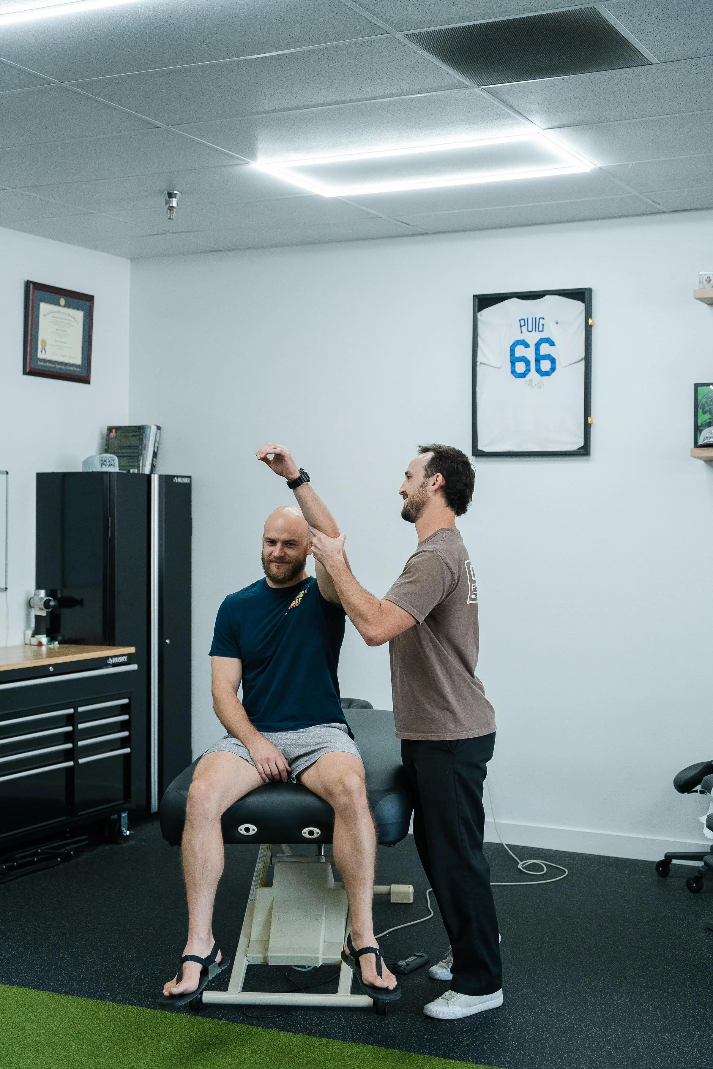 A man sitting on an examination table during a physical therapy session, with another man standing and applying a therapy technique to his arm, in a medical office with framed sports jerseys and certificates on the wall.