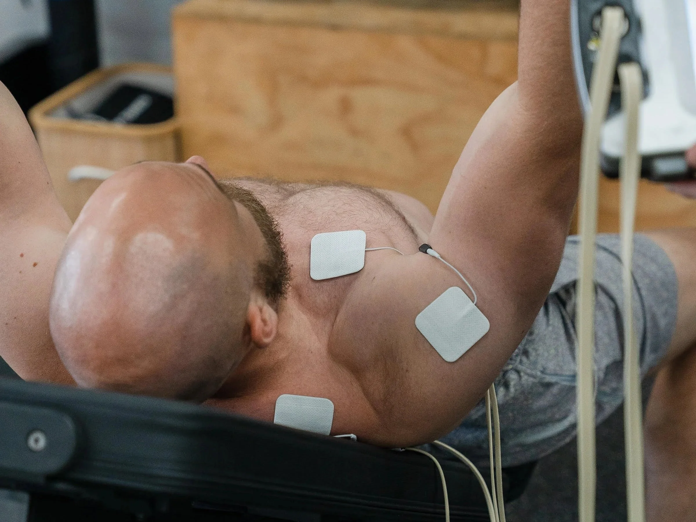 A man with a shaved head lying on his back in a medical setting with chest electrodes attached. He is wearing earphones and has a device placed on his lower abdomen, with wires connected to his chest.