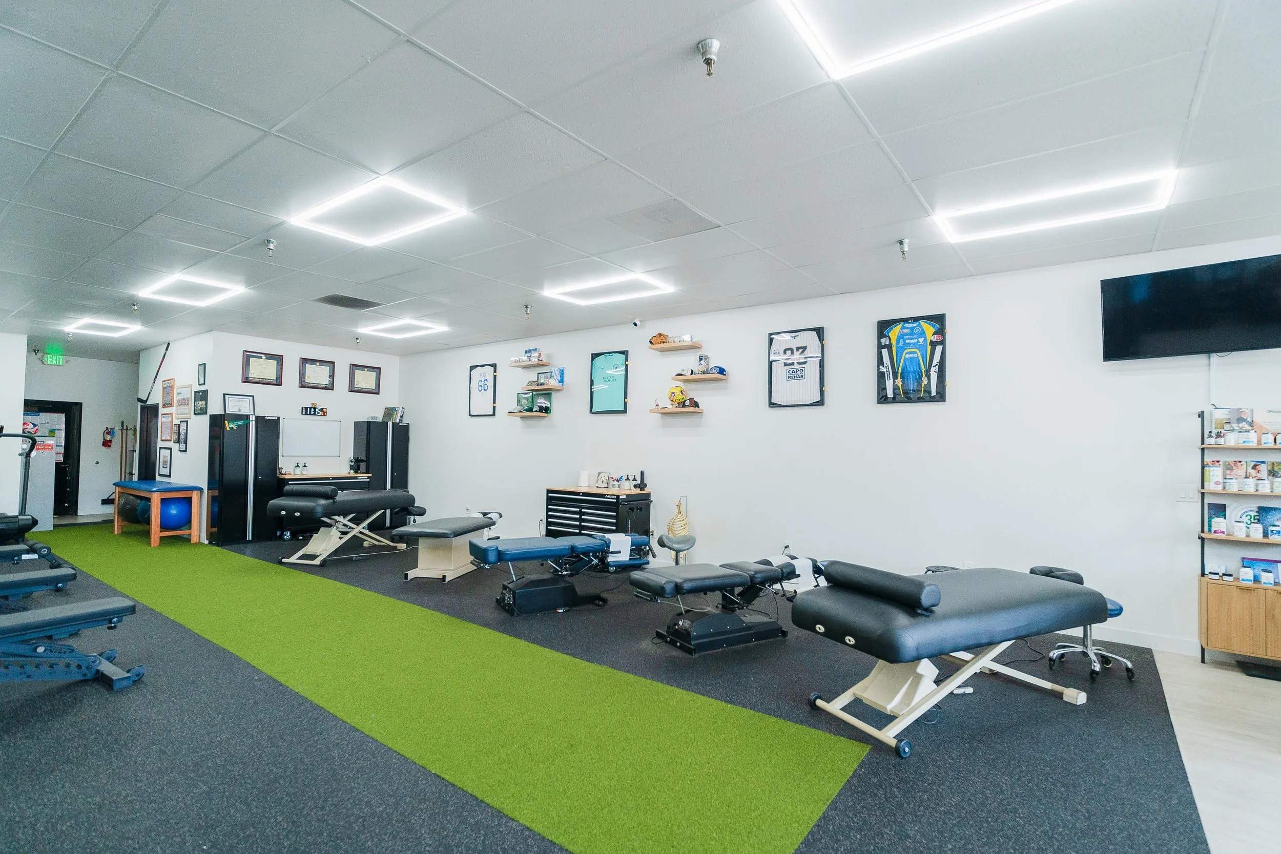 Indoor physical therapy or fitness room with exercise tables, equipment, wall decorations, and a television.