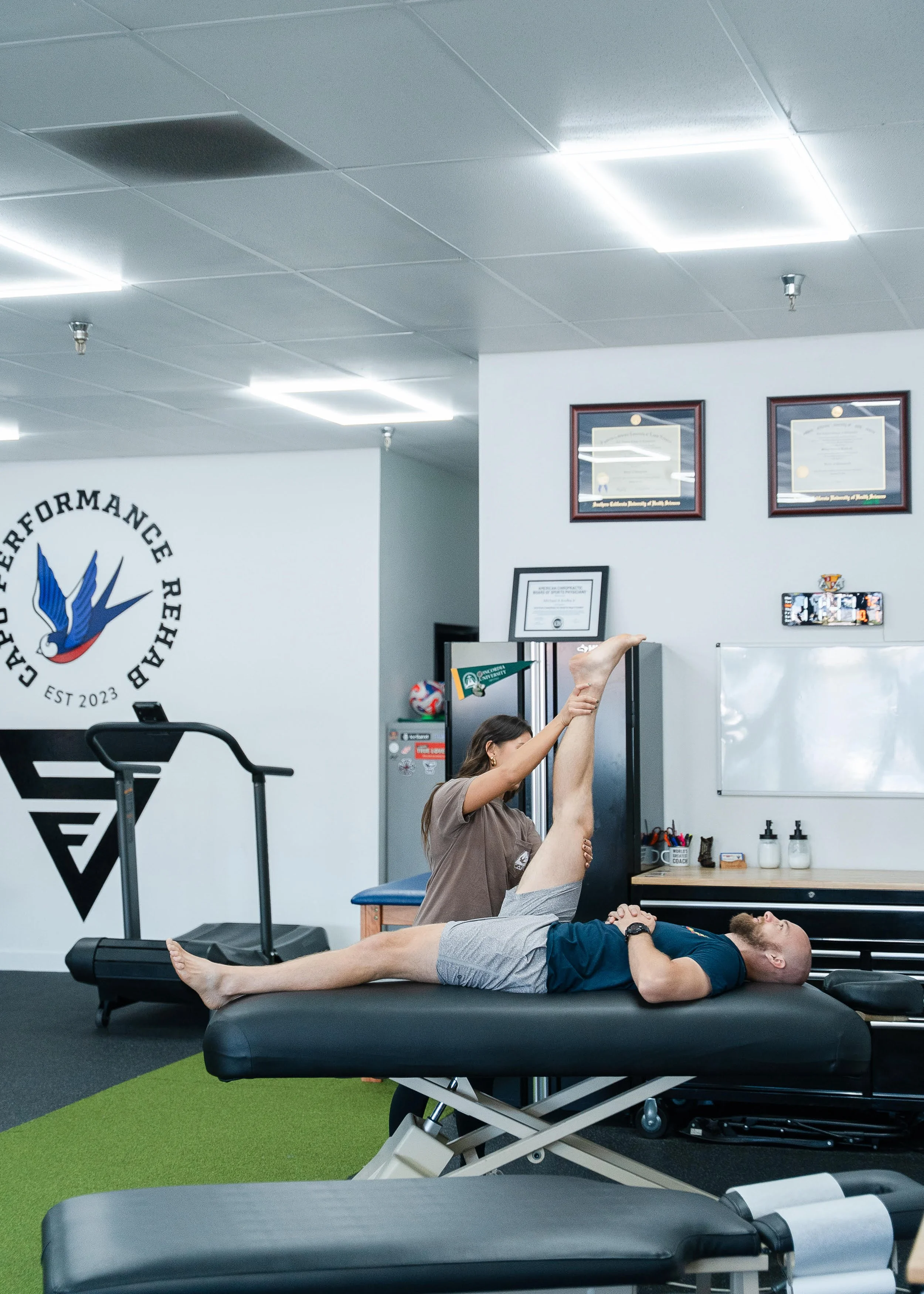 A person lying on a treatment table while another person stretches their leg in a physical therapy or fitness center. The room has framed certificates on the wall, a logo on a white wall, and various fitness equipment and supplies visible.