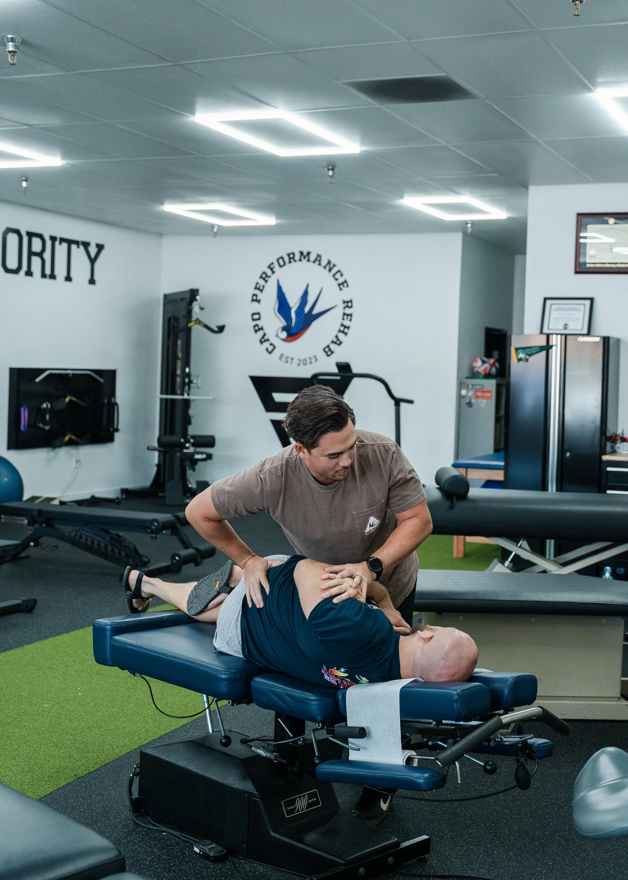 A chiropractor or physical therapist provides treatment to a patient lying face-up on a chiropractic table in a gym or therapy room. The background includes workout equipment and Capo Performance and Stronyx logos on the wall.