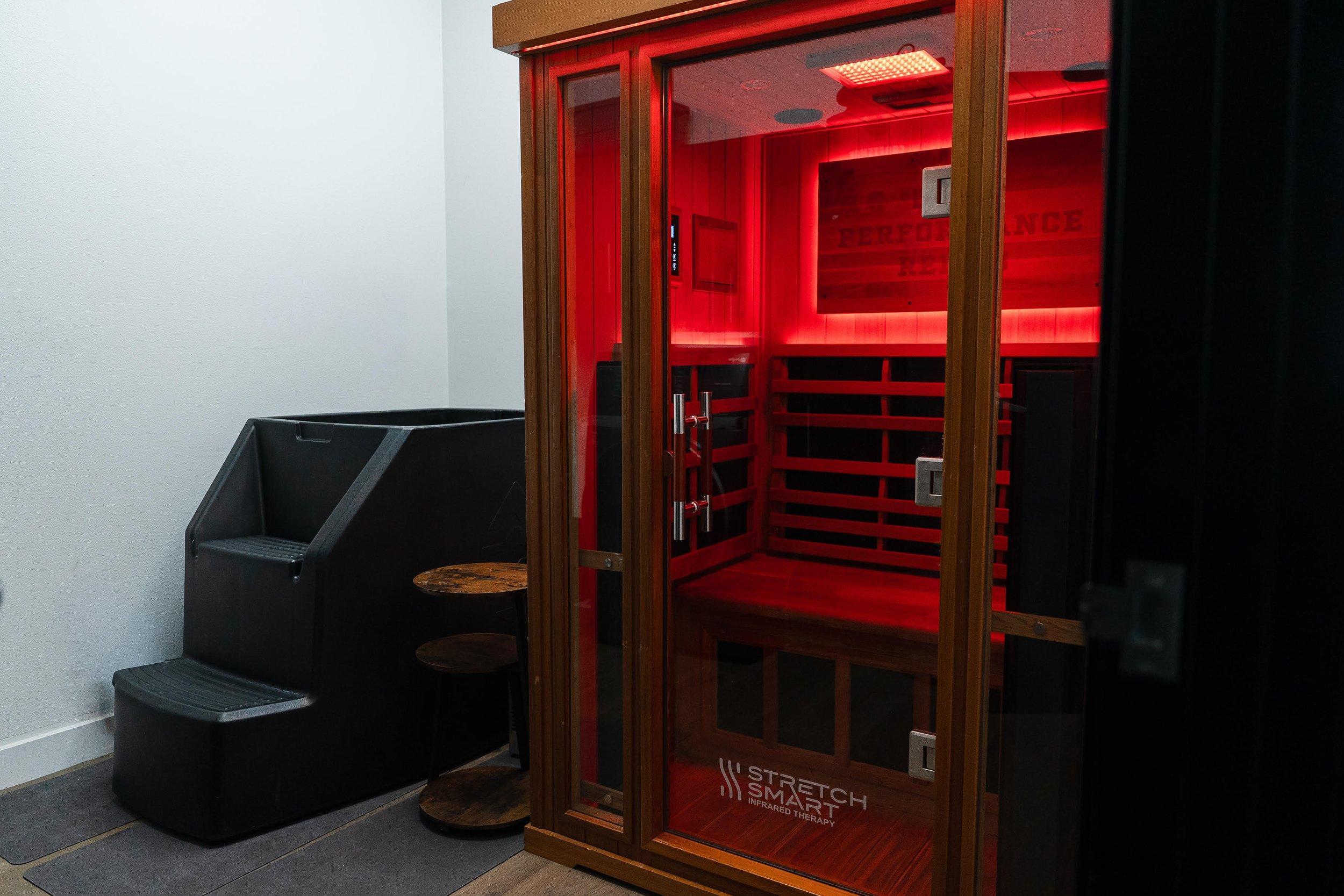 Infrared sauna cabinet with a red-glowing interior and a wooden exterior, next to a black plastic lounge chair with a small round table.