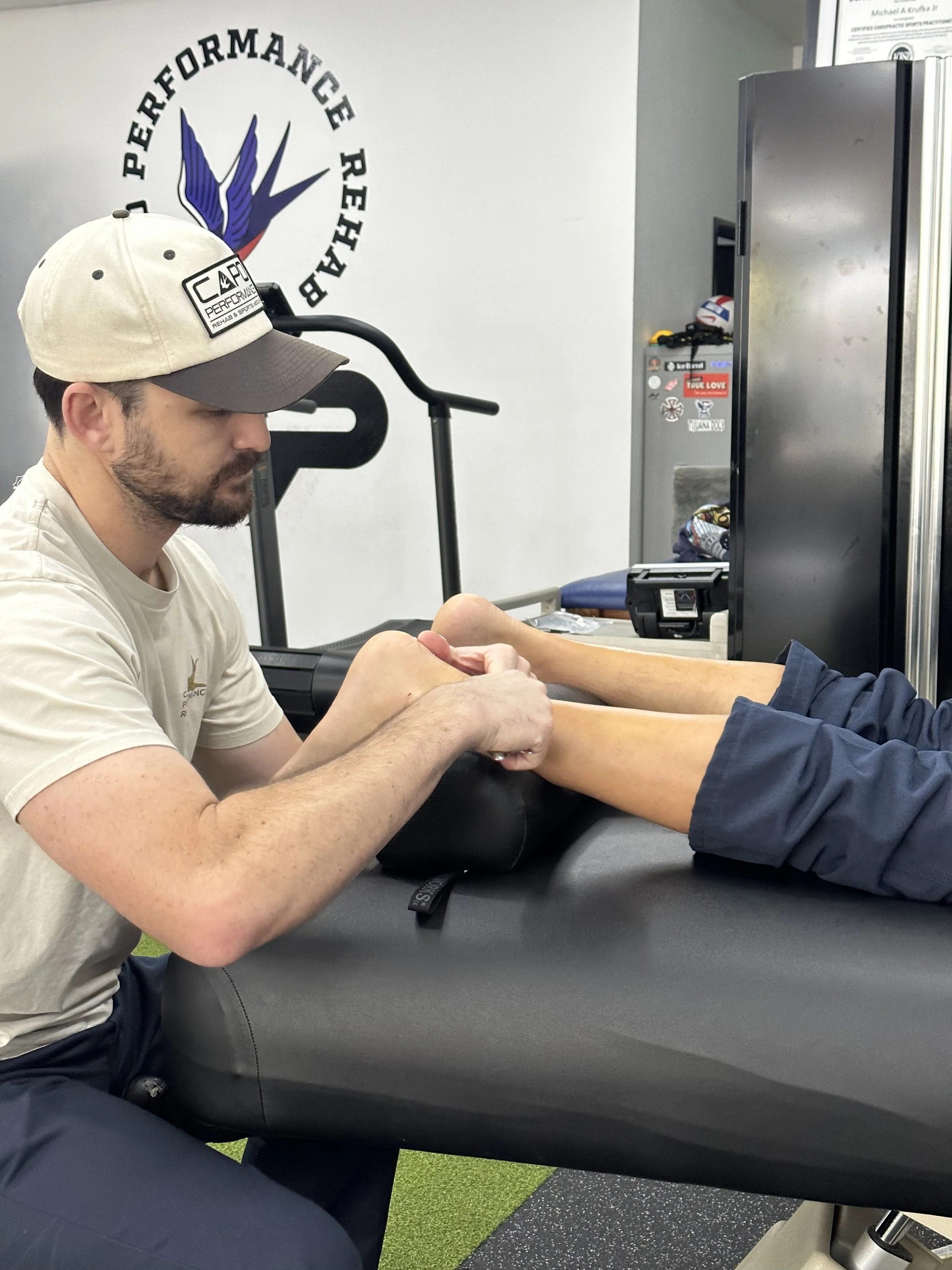 A physical therapist or trainer assisting a person with leg exercises in a therapy or gym setting. The background features a logo on the wall that reads 'Performance Rehab' with an abstract winged figure.