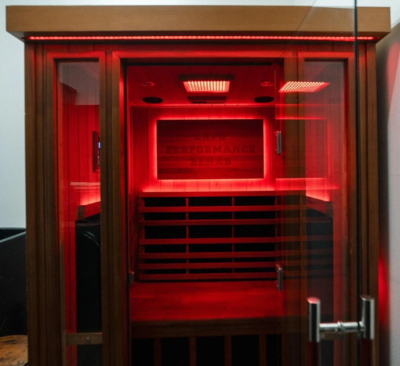 A wooden sauna with glass doors showcasing a red LED inside and a sign that reads 'Capo Performance.'