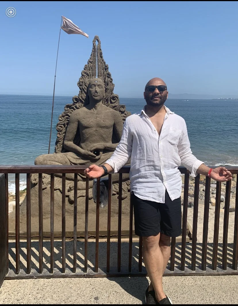 A Black man in a white button-down shirt standing in front of a buddha statue made of sand, in front of the ocean.