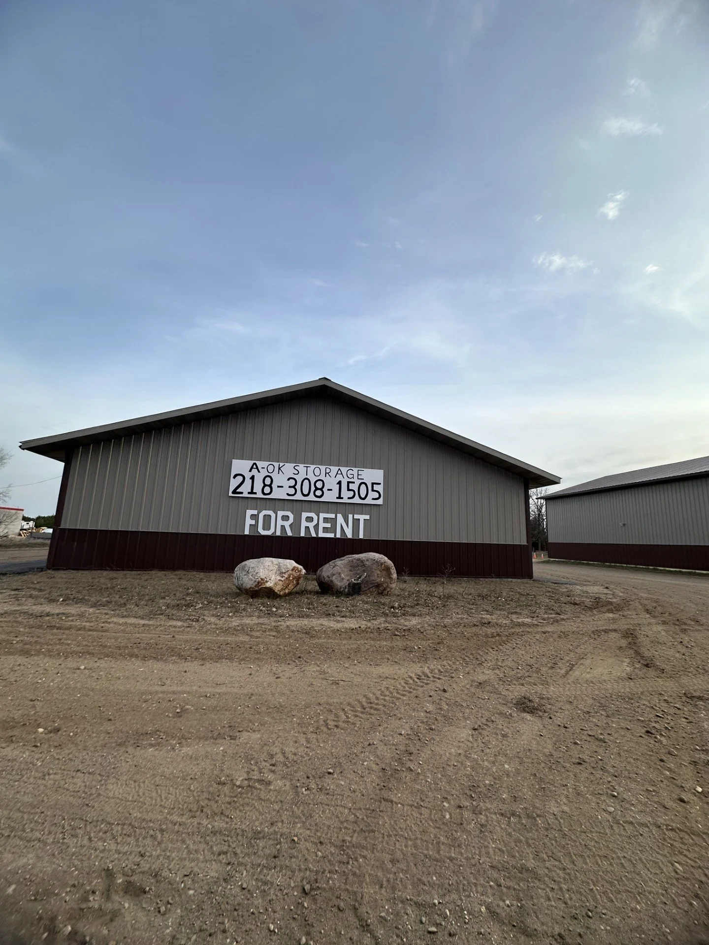 A large storage building with a sign that reads 'A-OK Storage 218-308-1505 FOR RENT' on its side, situated on a dirt lot with three rocks nearby under a partly cloudy sky.