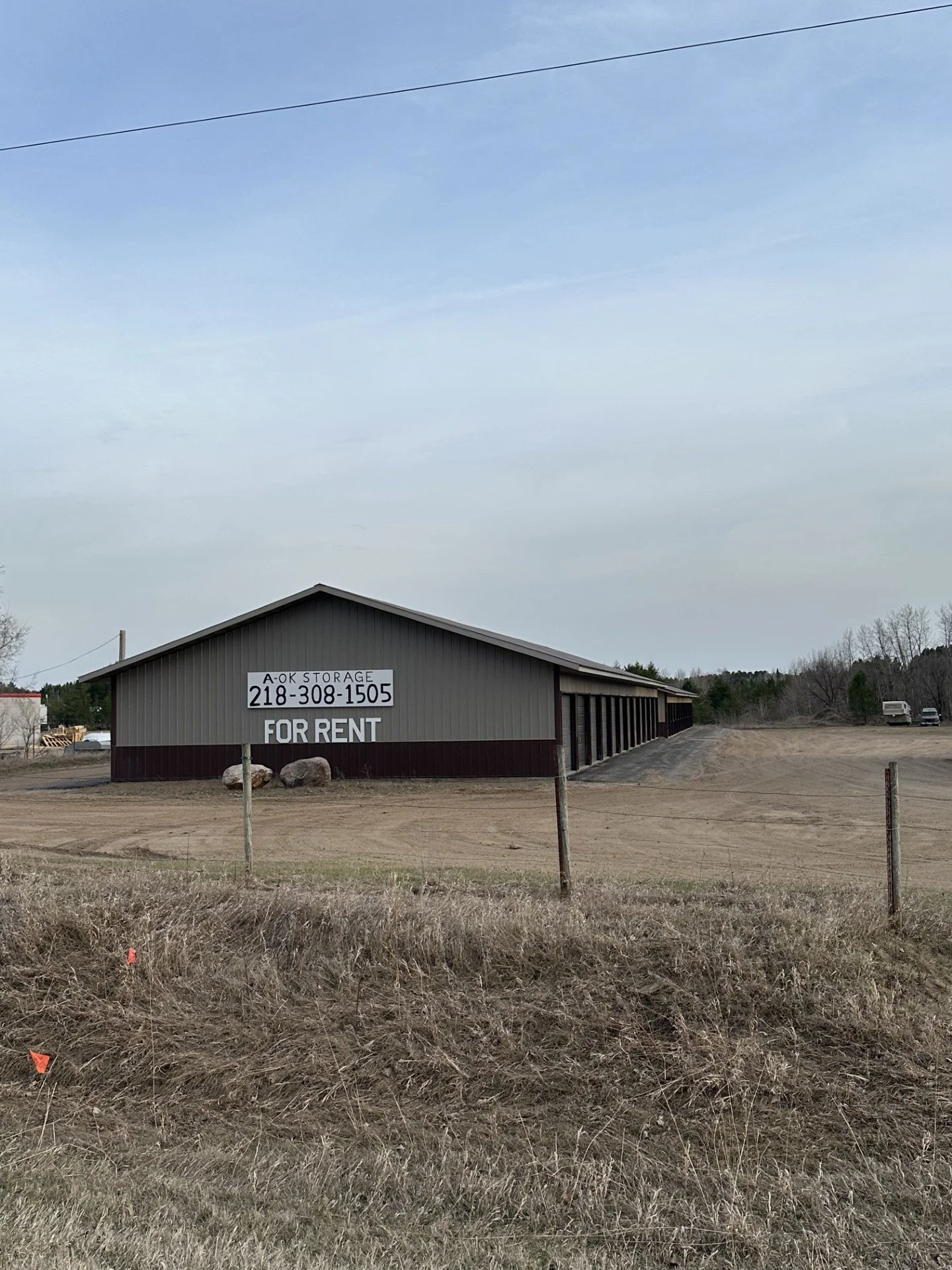 A large gray storage building with a sign that reads 'A-OK STORAGE 218-308-1505 FOR RENT' located on a dirt lot with some rocks and grass in the foreground, and trees and a vehicle in the background under a cloudy sky.
