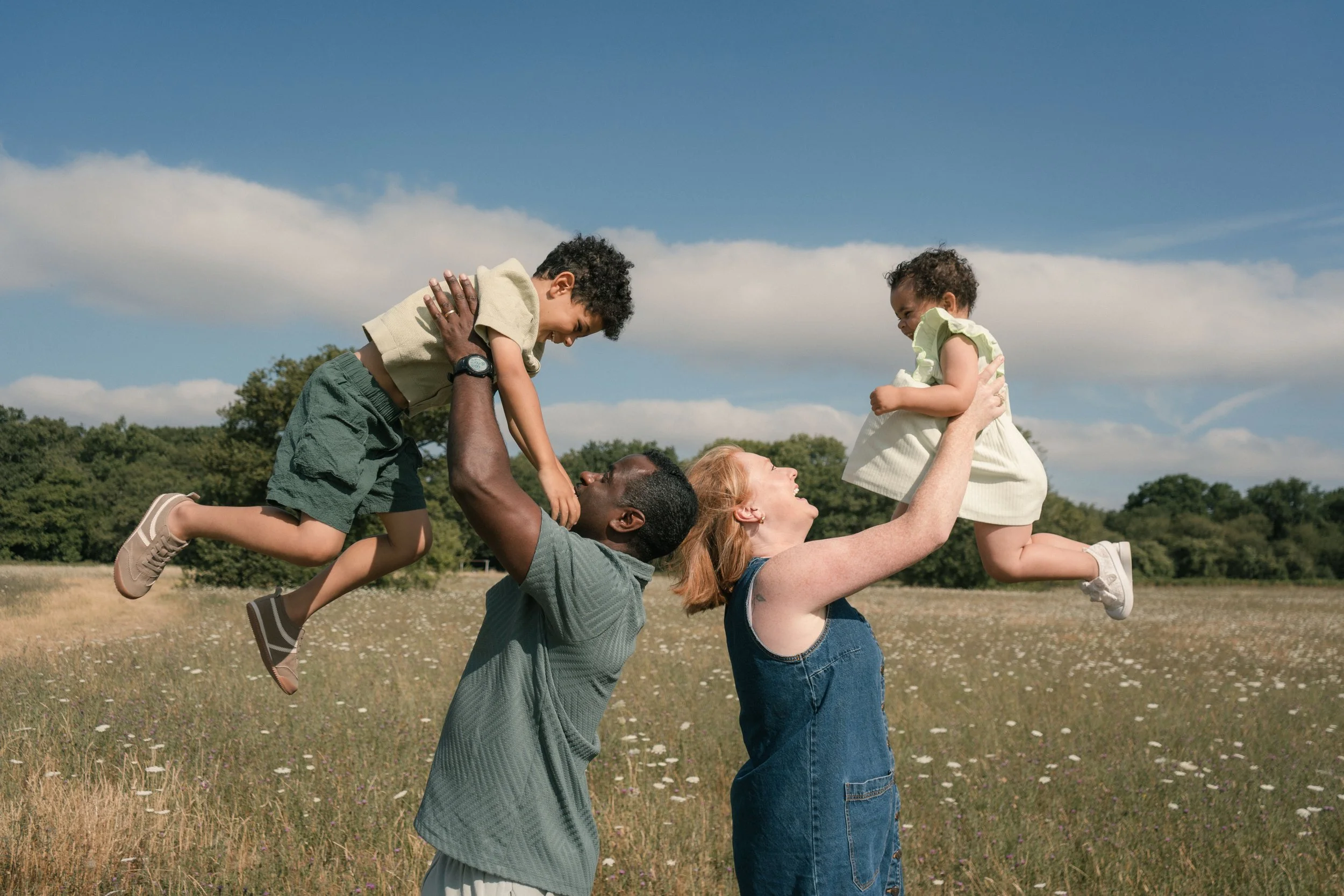 A group of four people playing in a field on a sunny day, with two adults holding up two children in the air, sharing a moment of joy.