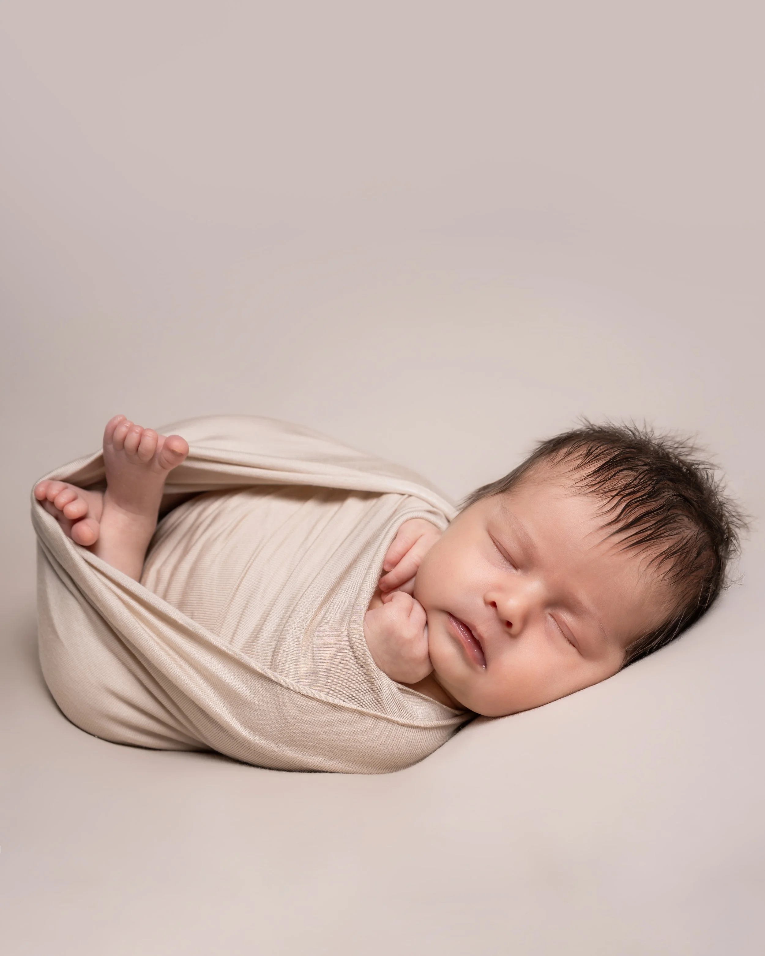 A sleeping baby with closed eyes, lying on a soft surface, wrapped in a beige swaddle blanket, with a neutral background.