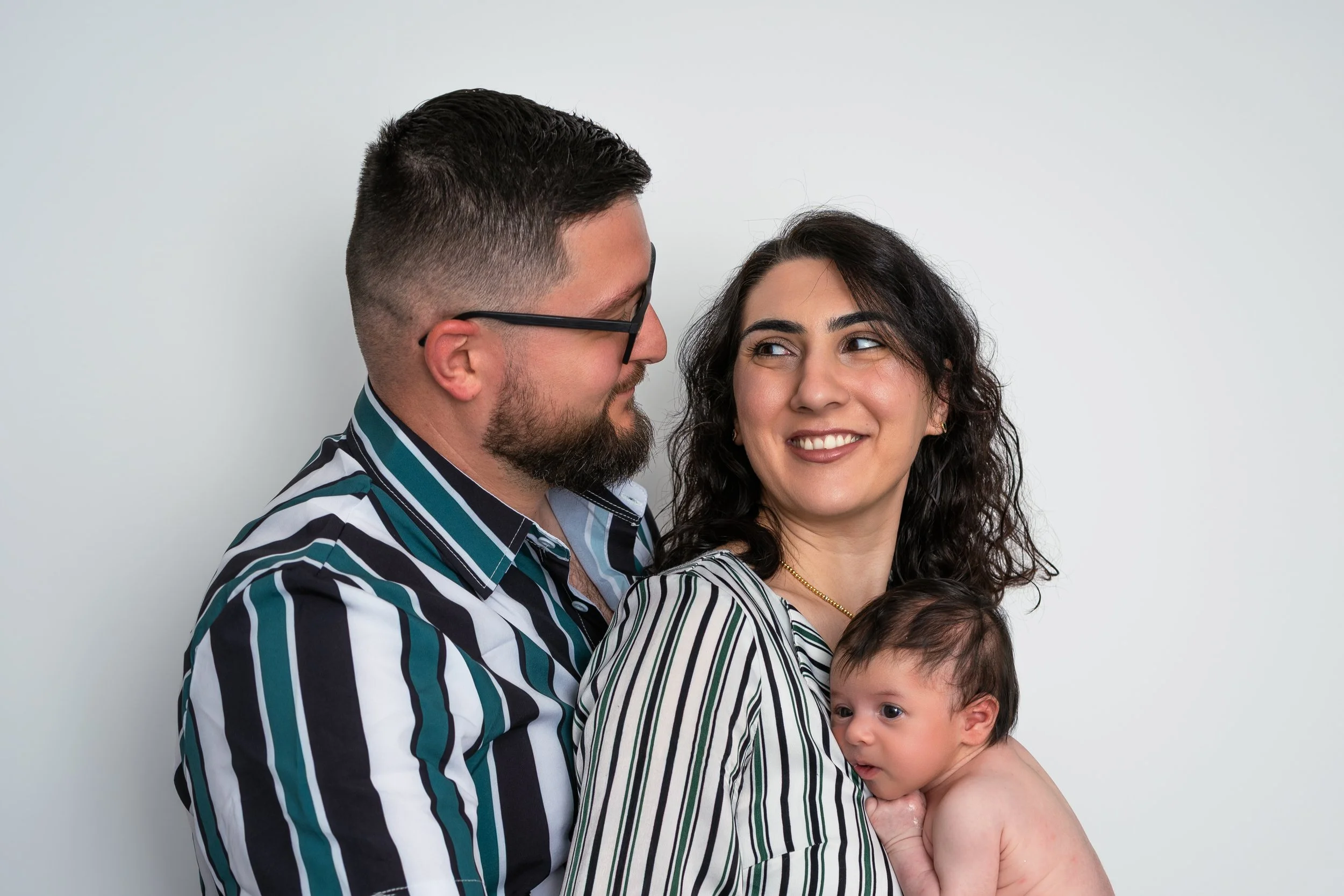 A man with glasses and a beard holds a young girl with dark curly hair and a striped shirt, while another child with light hair and no shirt is in front of them. They are standing against a plain white background.