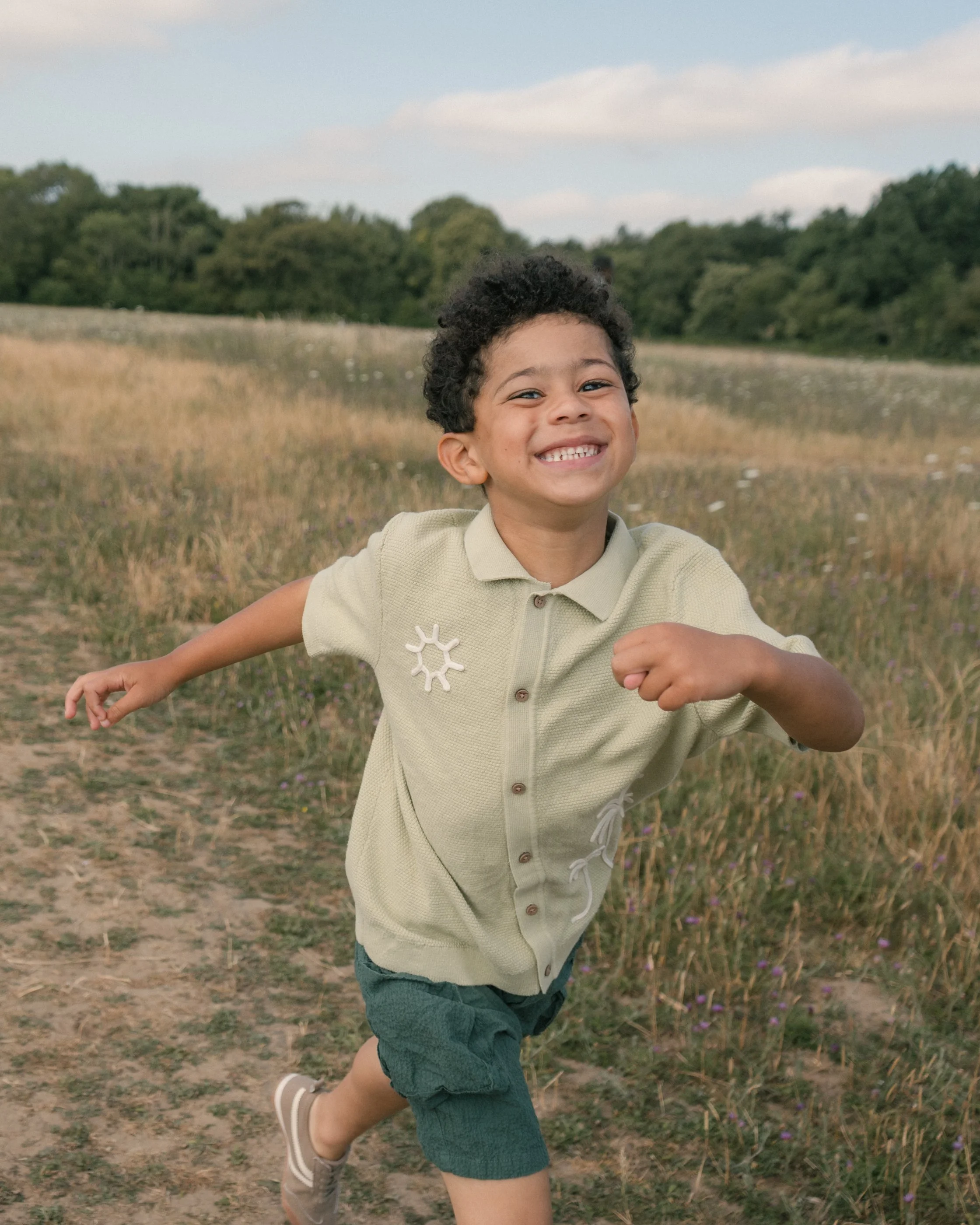 A young boy with curly hair and a big smile running through a field of tall grass and wildflowers, with trees and partly cloudy sky in the background.