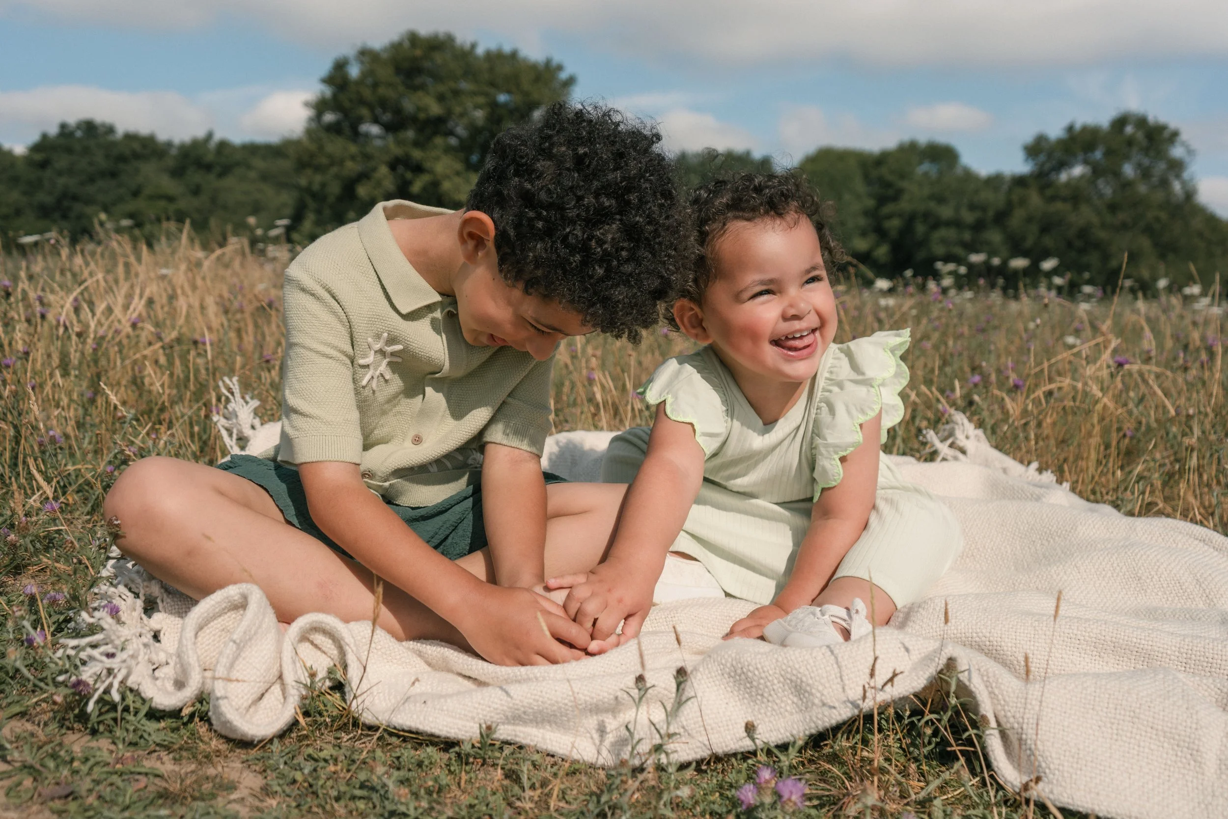 Two children, a boy and a girl, sitting on a blanket in a grassy field with wildflowers, laughing and playing together on a sunny day.