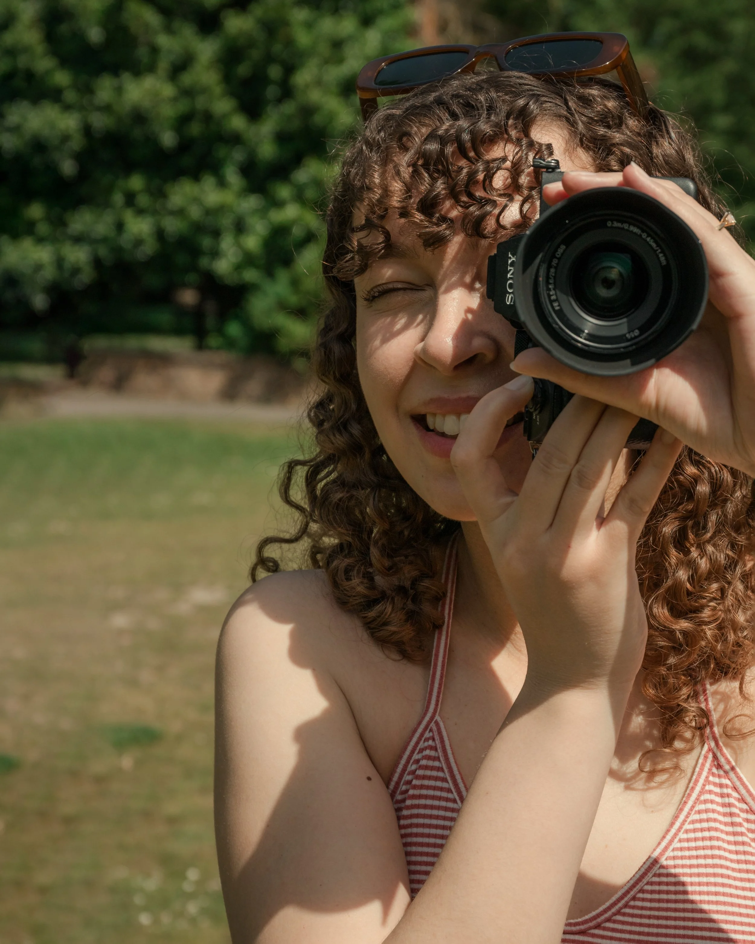 A woman with curly hair smiling, holding a camera up to her face for a photograph outdoors on a sunny day. She is wearing sunglasses on her head and a red and white striped top, with green trees and a park in the background.