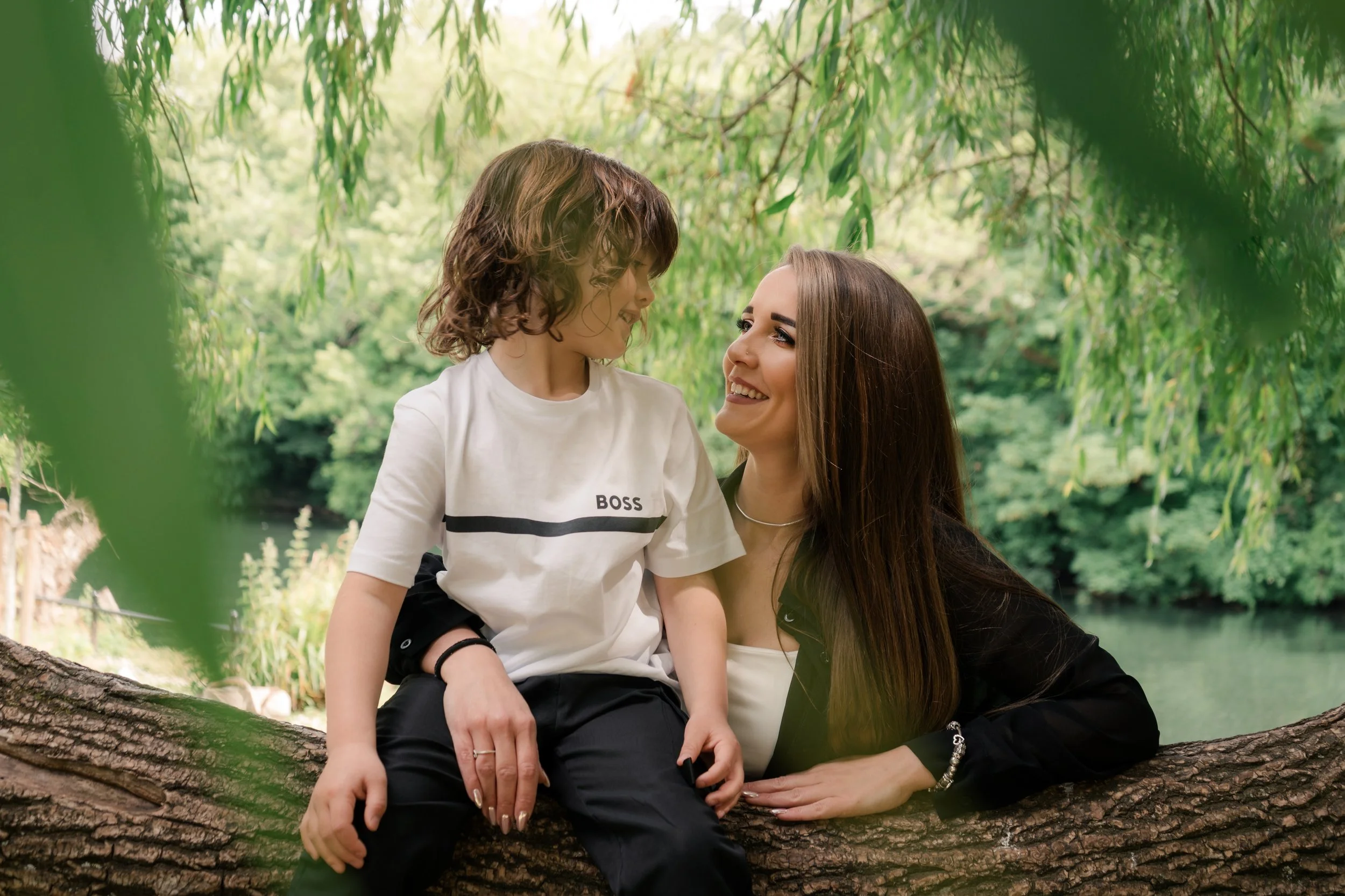 A woman and a young boy sitting on a tree branch outdoors, smiling at each other, surrounded by green foliage.