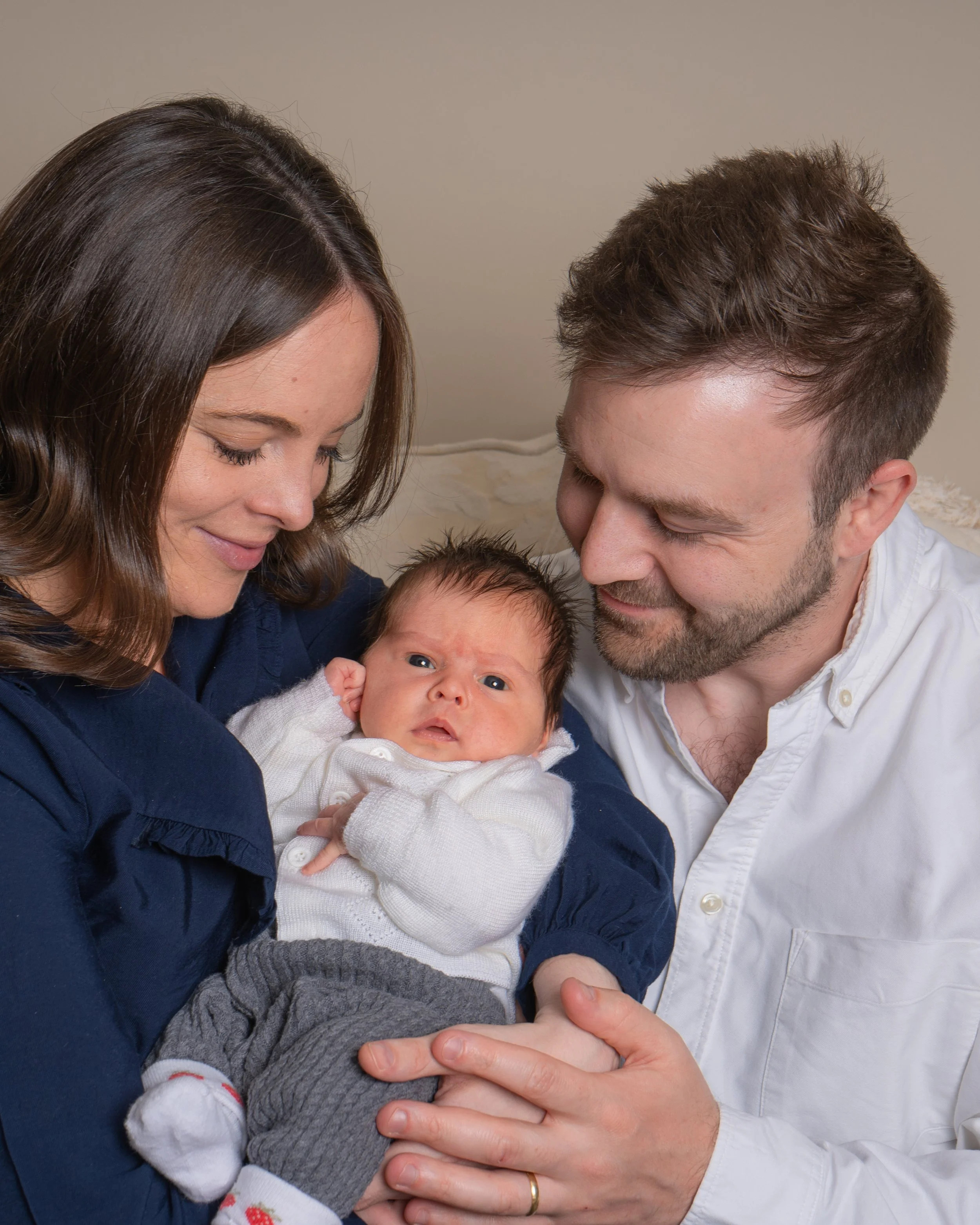 A family of three, including a woman, a man, and a baby, is close together and smiling softly, with the baby laying on the woman's lap. The woman has brown hair, and the man has short brown hair and facial stubble. The baby has dark hair and is wearing a white onesie with small patterns. The family appears affectionate and joyful.