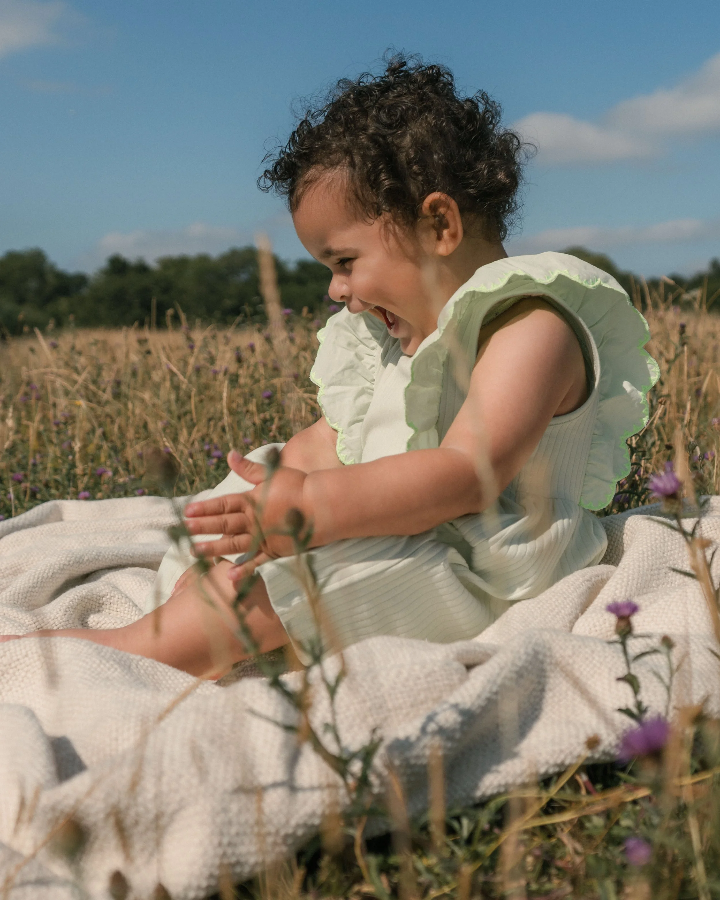 A young child with curly hair and a joyful expression sitting outdoors in a field of purple flowers under a blue sky.