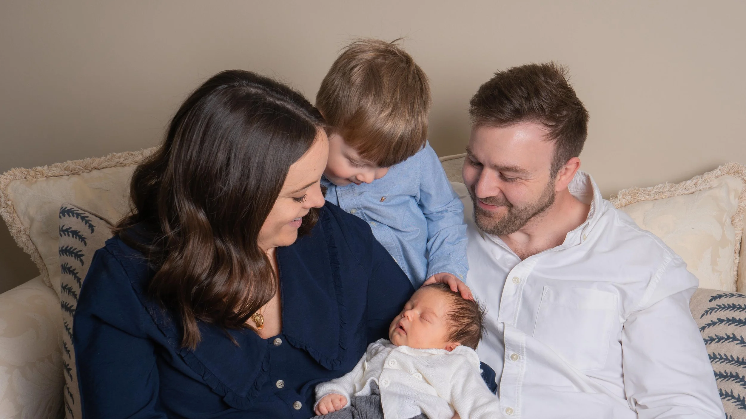Family of five, including two parents, two children, and an infant, sitting closely on a couch, smiling and interacting with each other, in a cozy home setting.