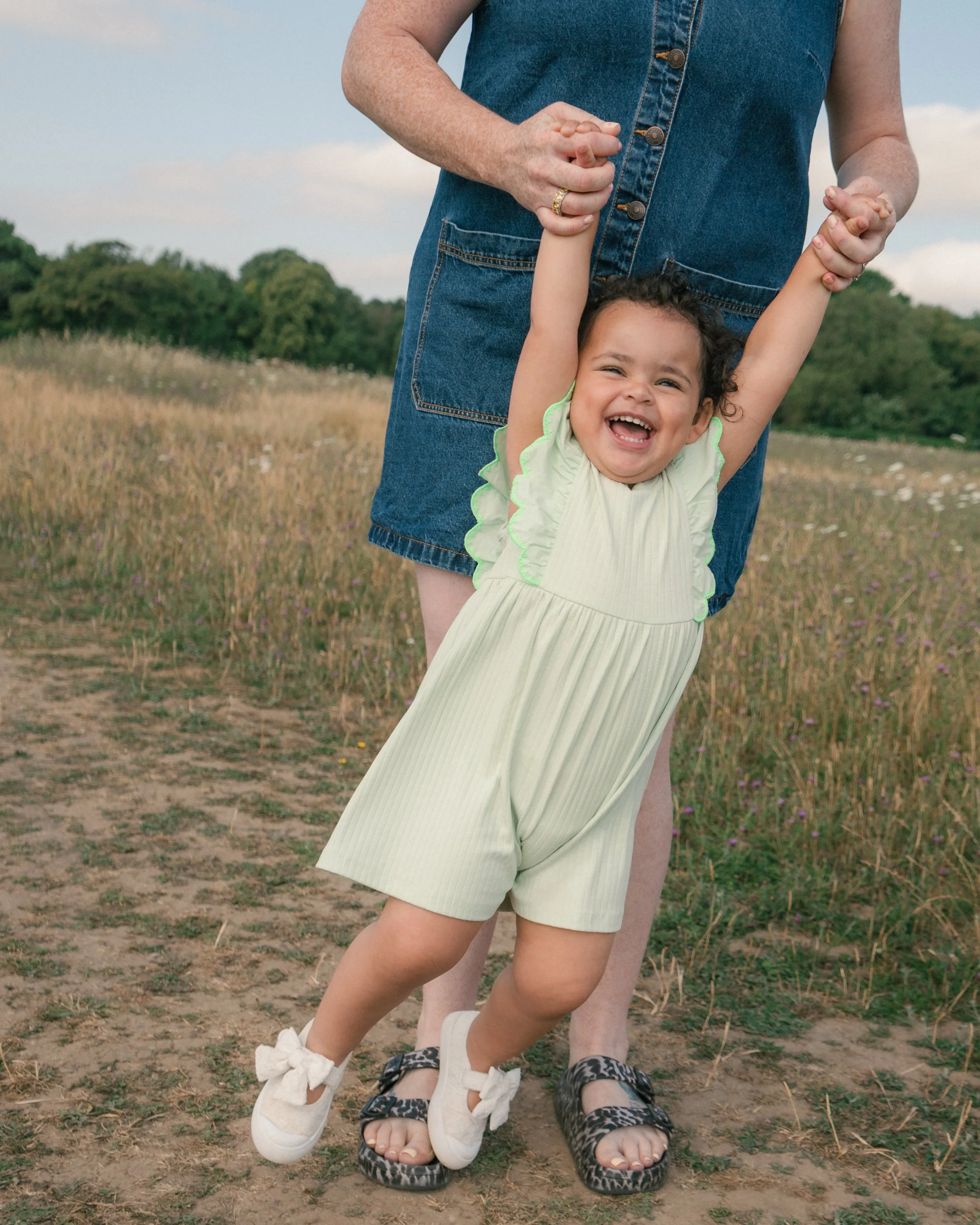 A young girl with curly hair and a big smile, wearing a yellow dress, holding hands with an adult, standing outdoors in a grassy field with trees in the background.