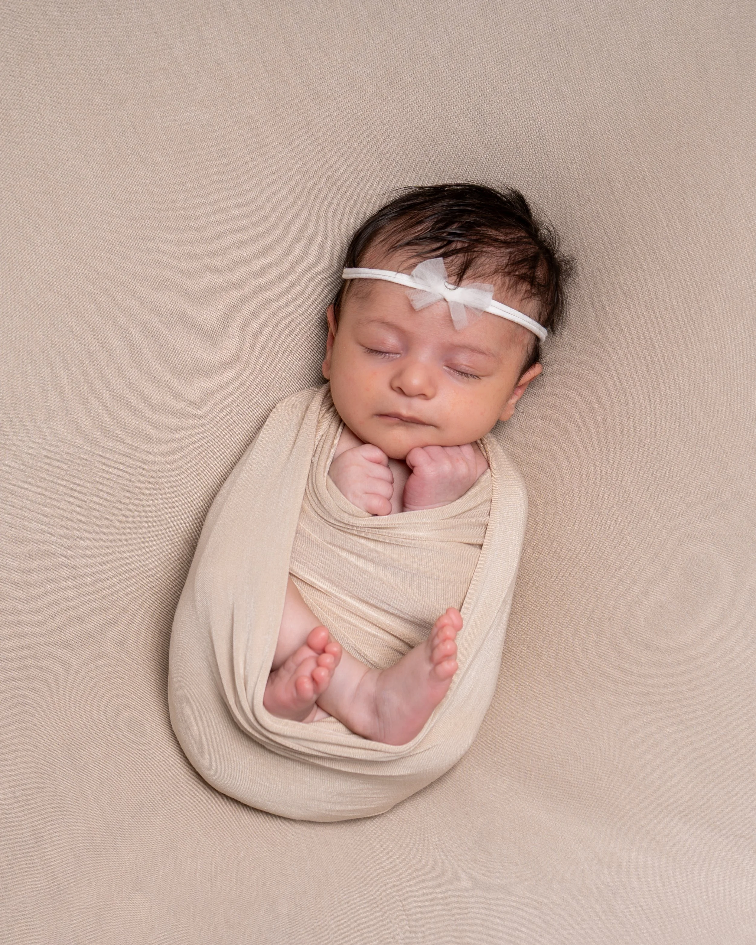 A sleeping baby with dark, wet hair wrapped in a beige blanket on a beige background, wearing a white headband with a bow.