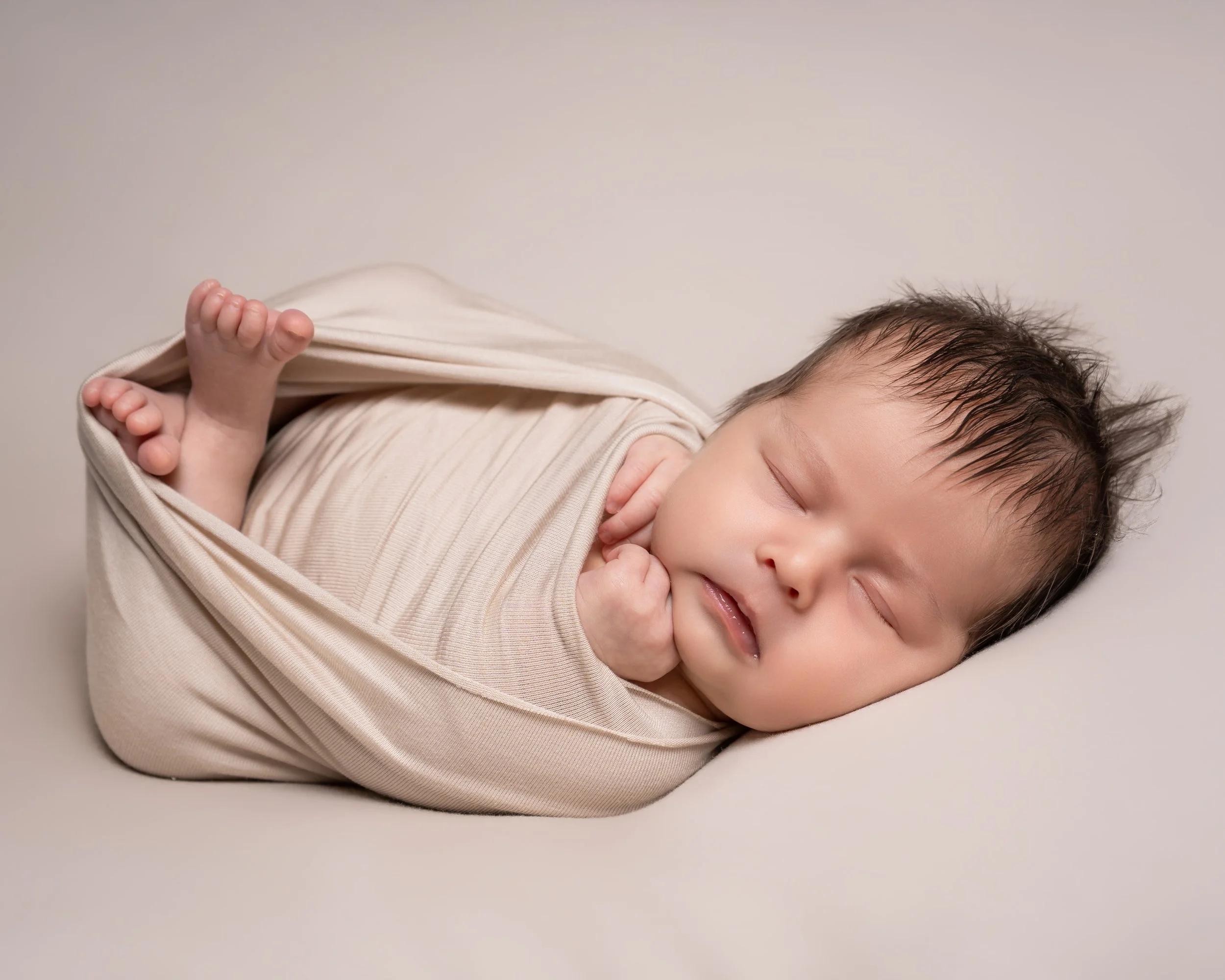 A sleeping baby wrapped in a beige blanket against a plain light-colored background.