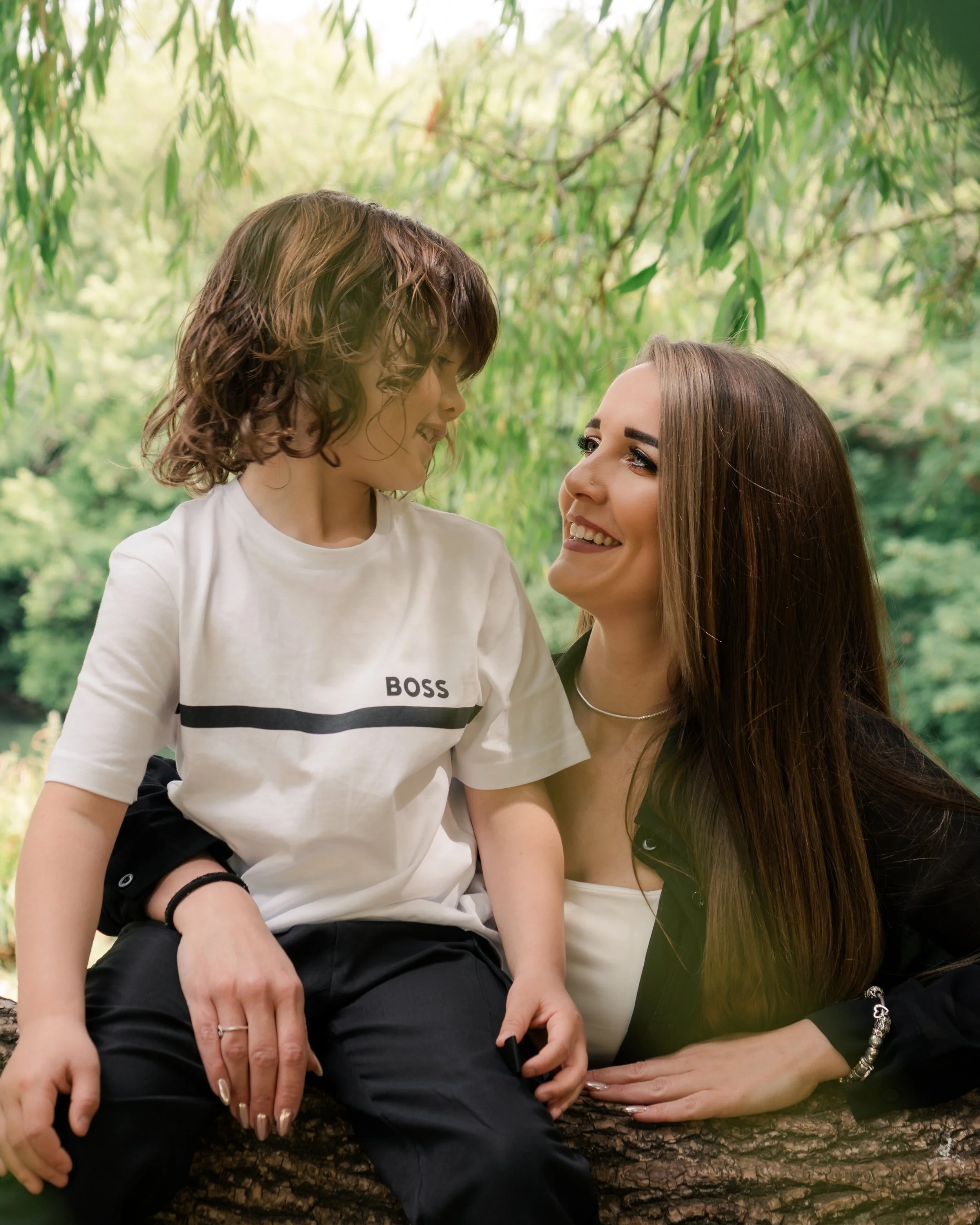 A smiling woman with long brunette hair, wearing a white top, is sitting or leaning next to a young boy with curly brown hair and a white t-shirt, in a lush, green outdoor setting with trees in the background.