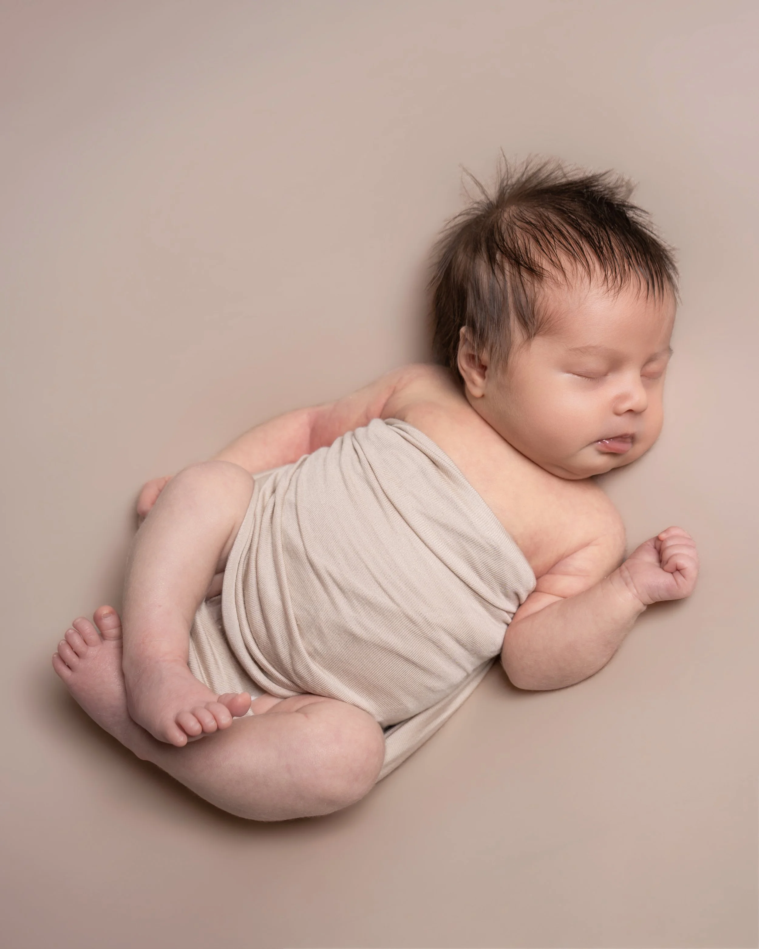 A baby with closed eyes, light skin, and dark hair lying on a beige background, wearing a beige onesie, with an adult's hand gently supporting the baby's back.
